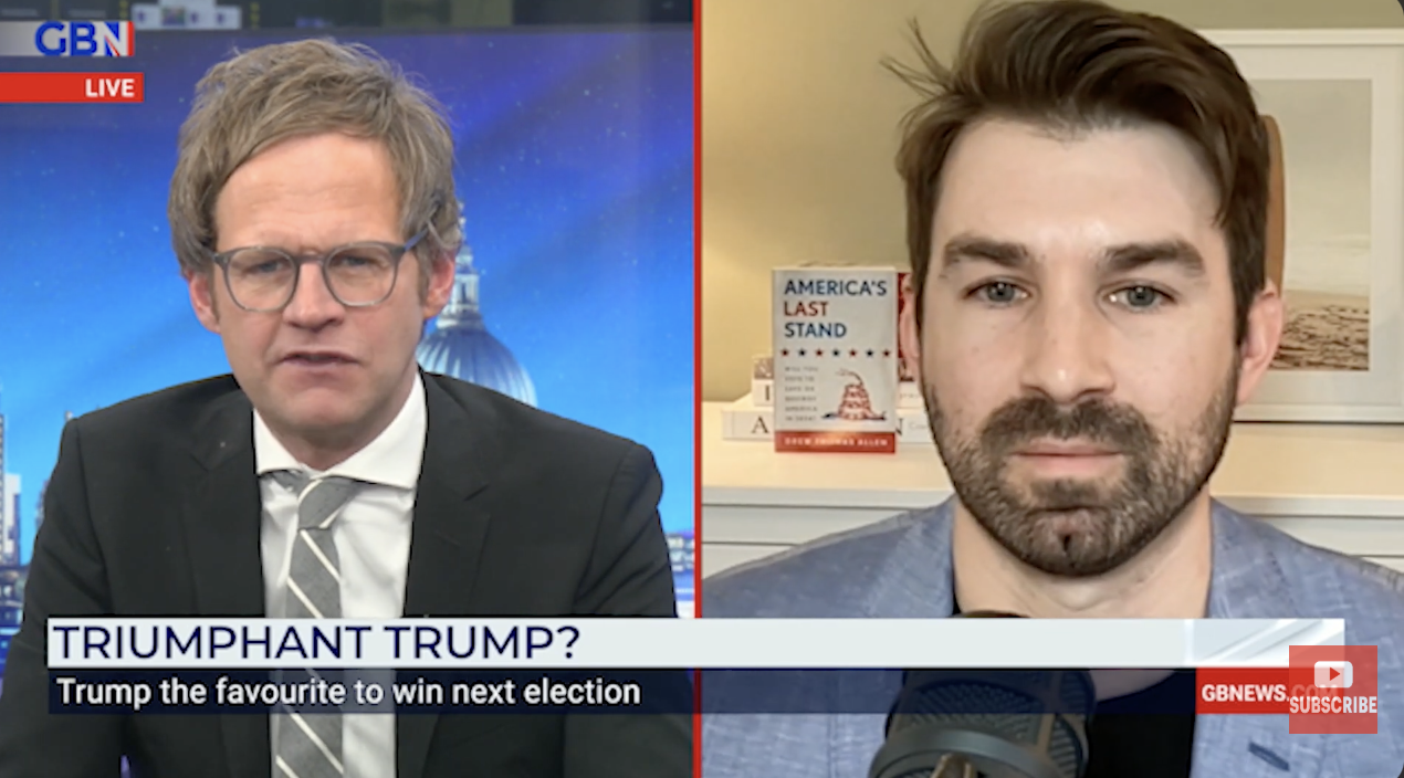 Screenshot of a GB News broadcast with two male commentators discussing the possibility of Trump winning the next election. The left commentator has glasses, curly light brown hair, and is wearing a black suit with a striped shirt and a striped tie. The right commentator has dark brown hair, a beard, and is wearing a light blue blazer over a dark shirt. In the background, the left has a cityscape with a prominent government building, and the right has a bookshelf with a book titled 'America's Last Stand' and a framed photograph.