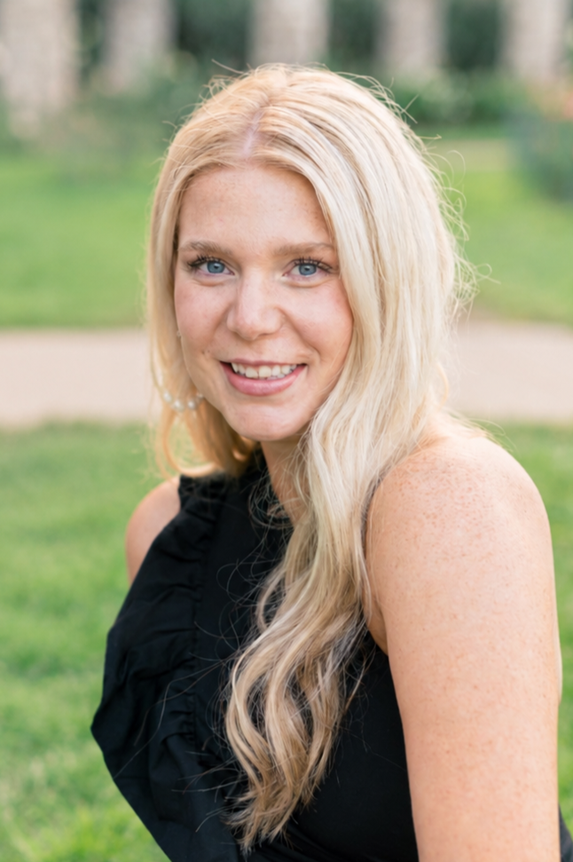 A young woman with long blonde hair, blue eyes, and freckles, smiling outdoors with green grass and trees in the background, wearing a sleeveless black top.