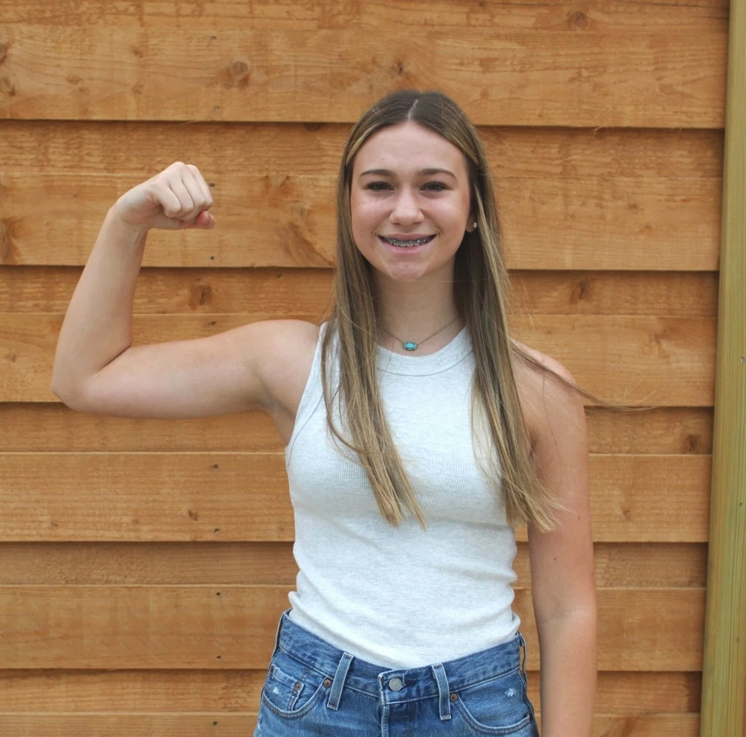 Teen girl with long hair smiling and flexing her right arm, wearing a white tank top and jeans, standing in front of a wooden wall.