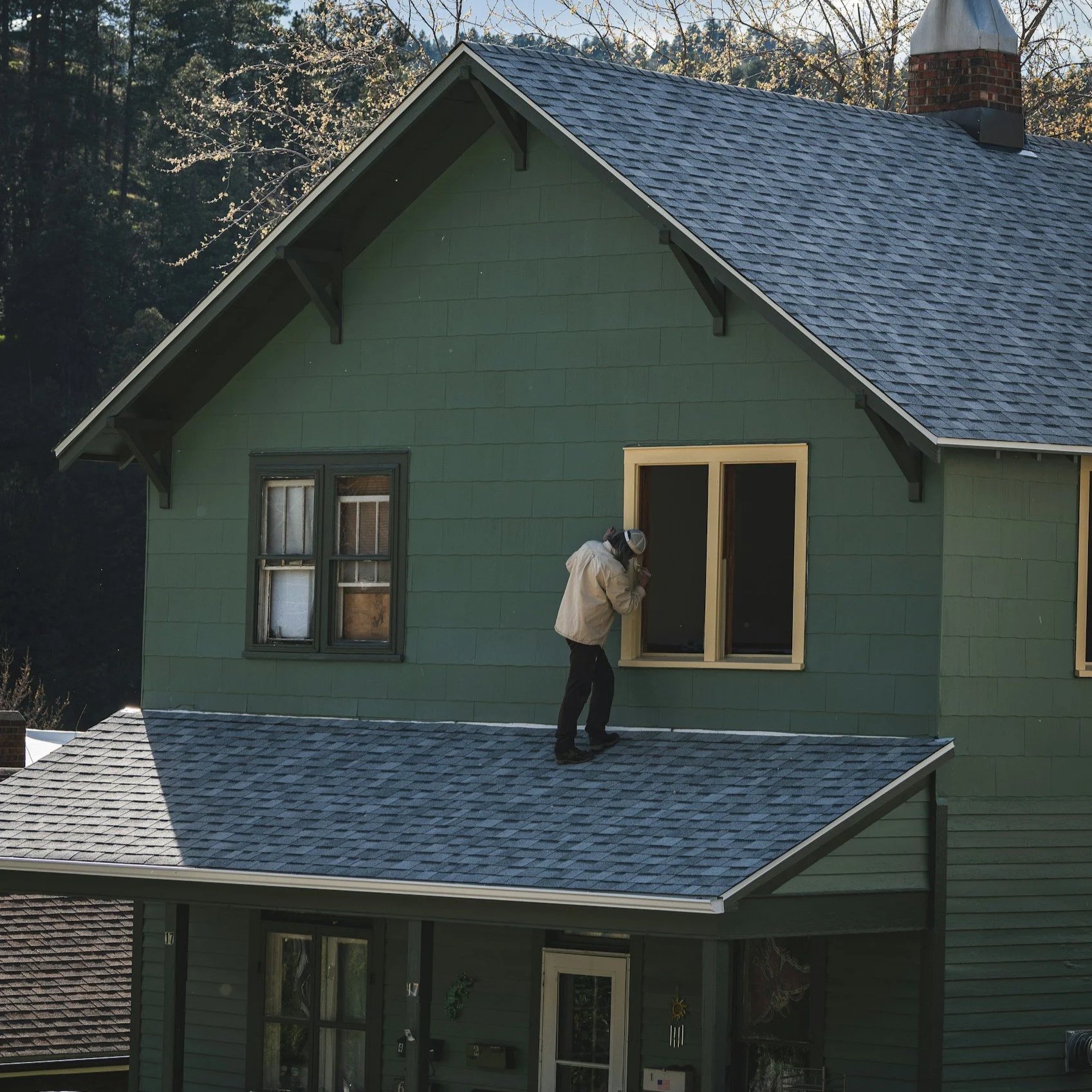 A person in protective gear working on the exterior of a house with green siding and blue shingle roof, surrounded by trees.
