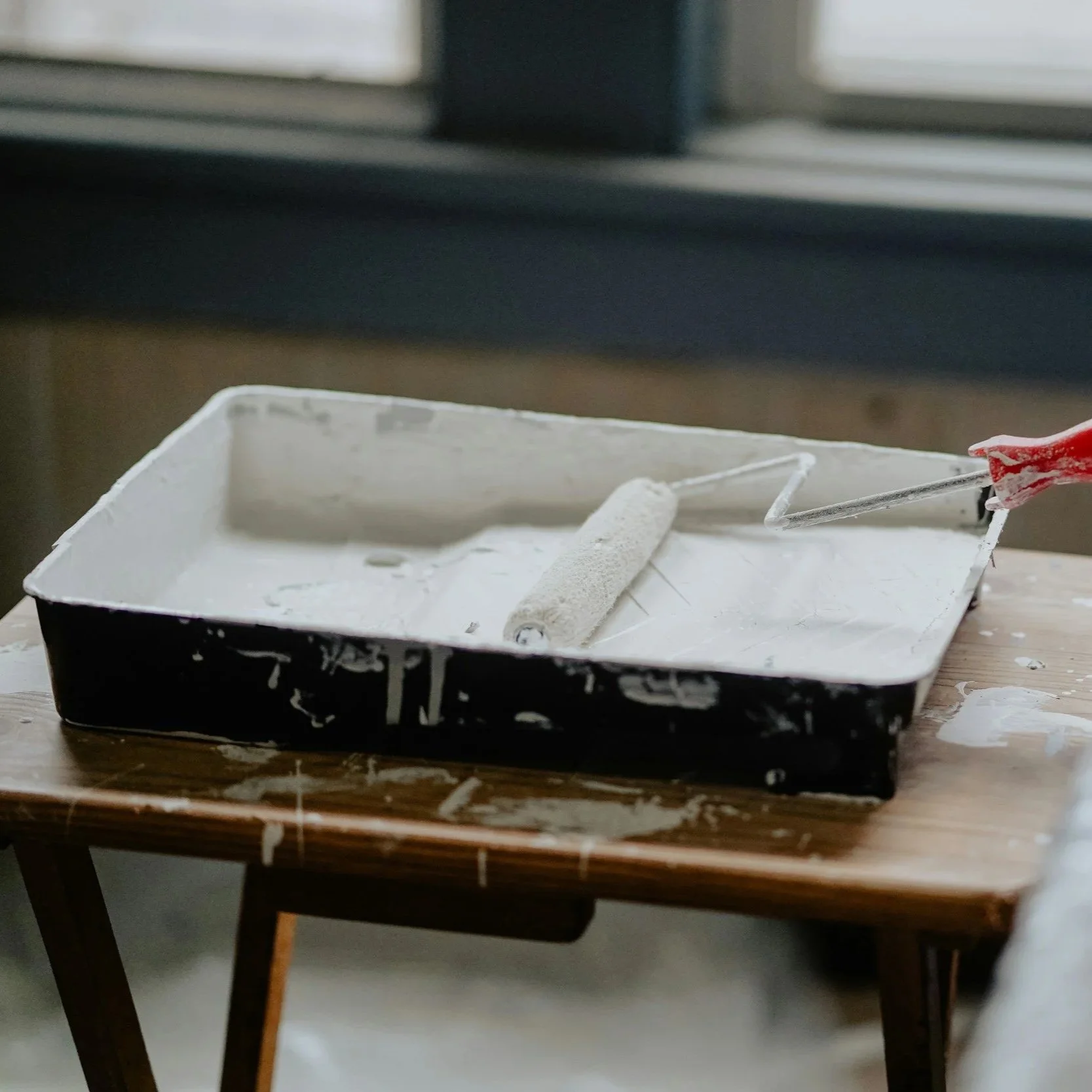 A paint roller and paint tray on a wooden table, with spilled white paint around the table.