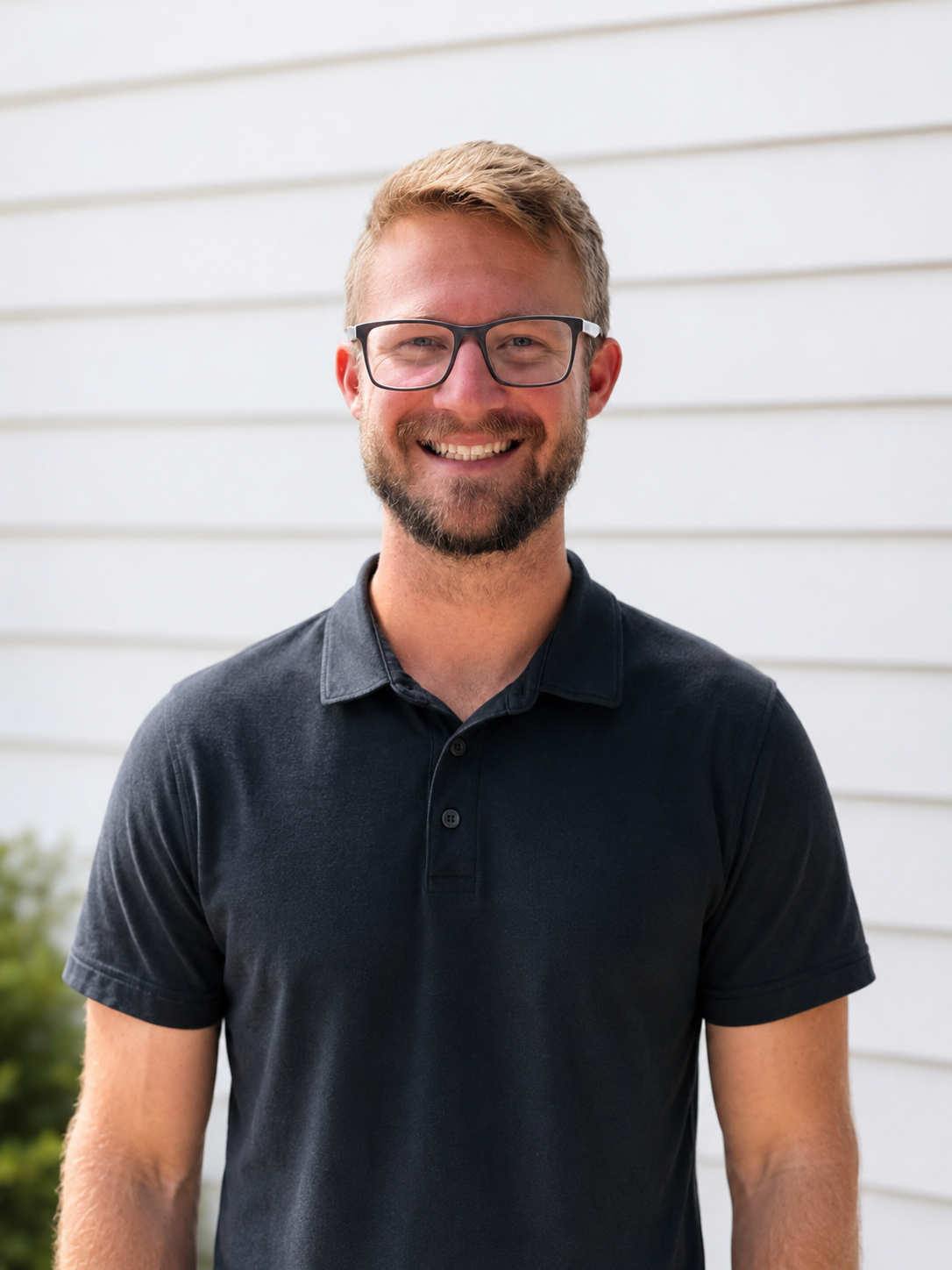 The business owner with glasses, a beard, and short blond hair stands outdoors in front of a white siding wall, wearing a black polo shirt.