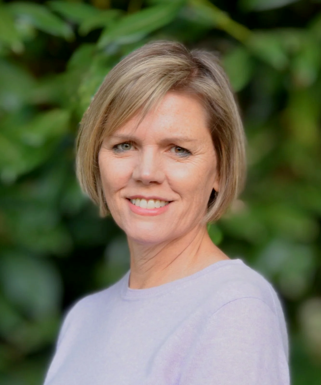 A woman with short blonde hair and a smile, wearing a light-colored shirt, standing outdoors with green foliage in the background.