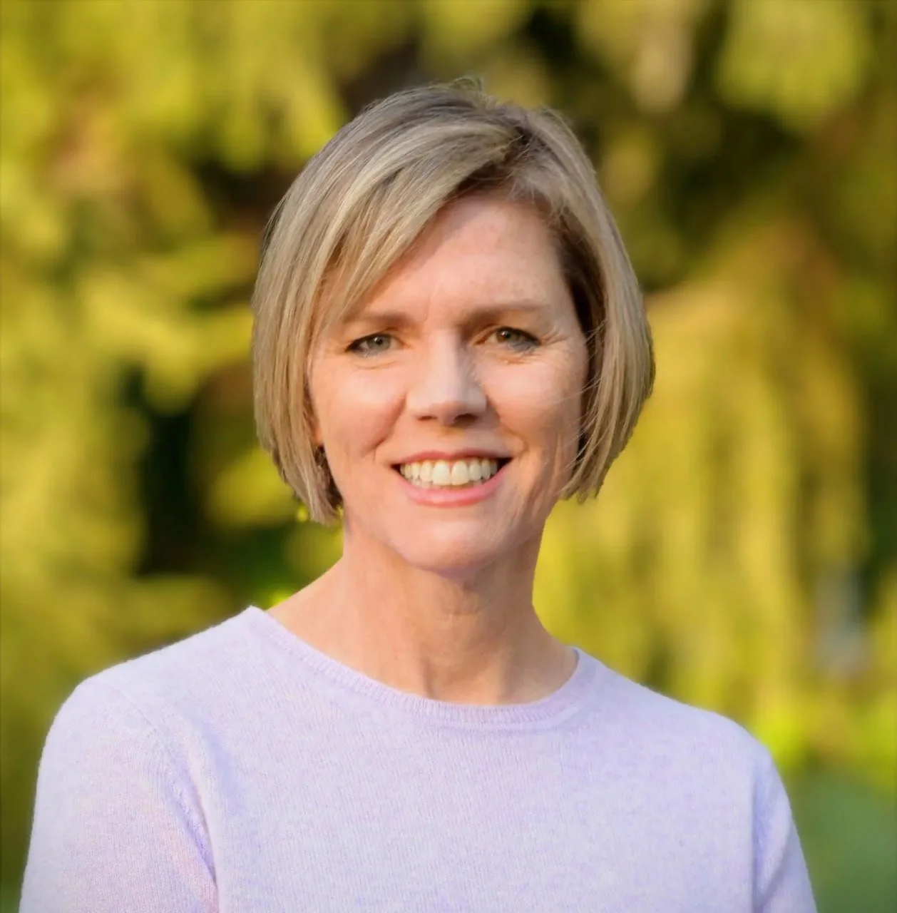 A woman with short blonde hair smiling outdoors with a blurred green and yellow background.