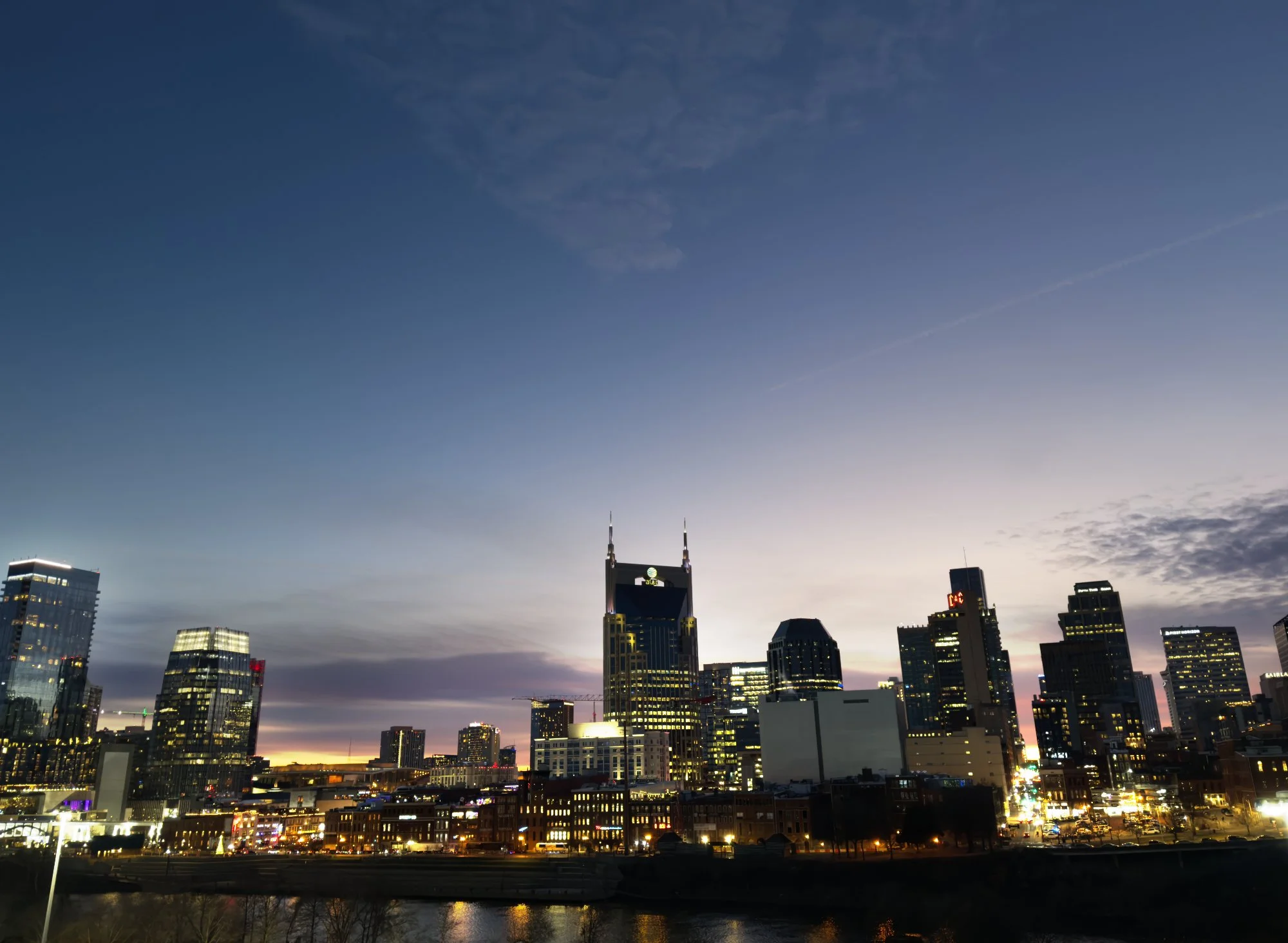 A city skyline at dusk featuring tall skyscrapers with illuminated windows and a few clouds in the sky. The city lights reflect on the water in the foreground.