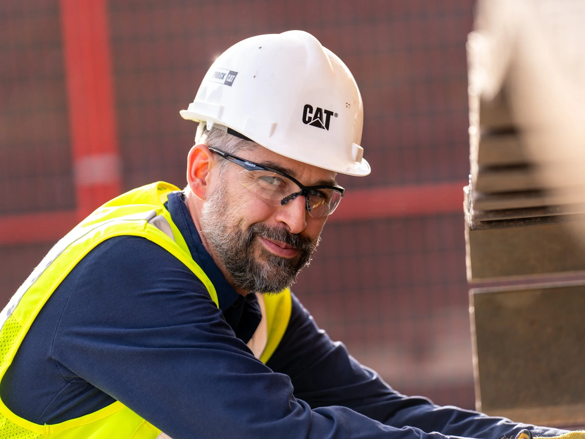 A construction worker with glasses and a beard, wearing a white safety helmet with CAT logo and a yellow reflective vest, smiling while working on a construction site.