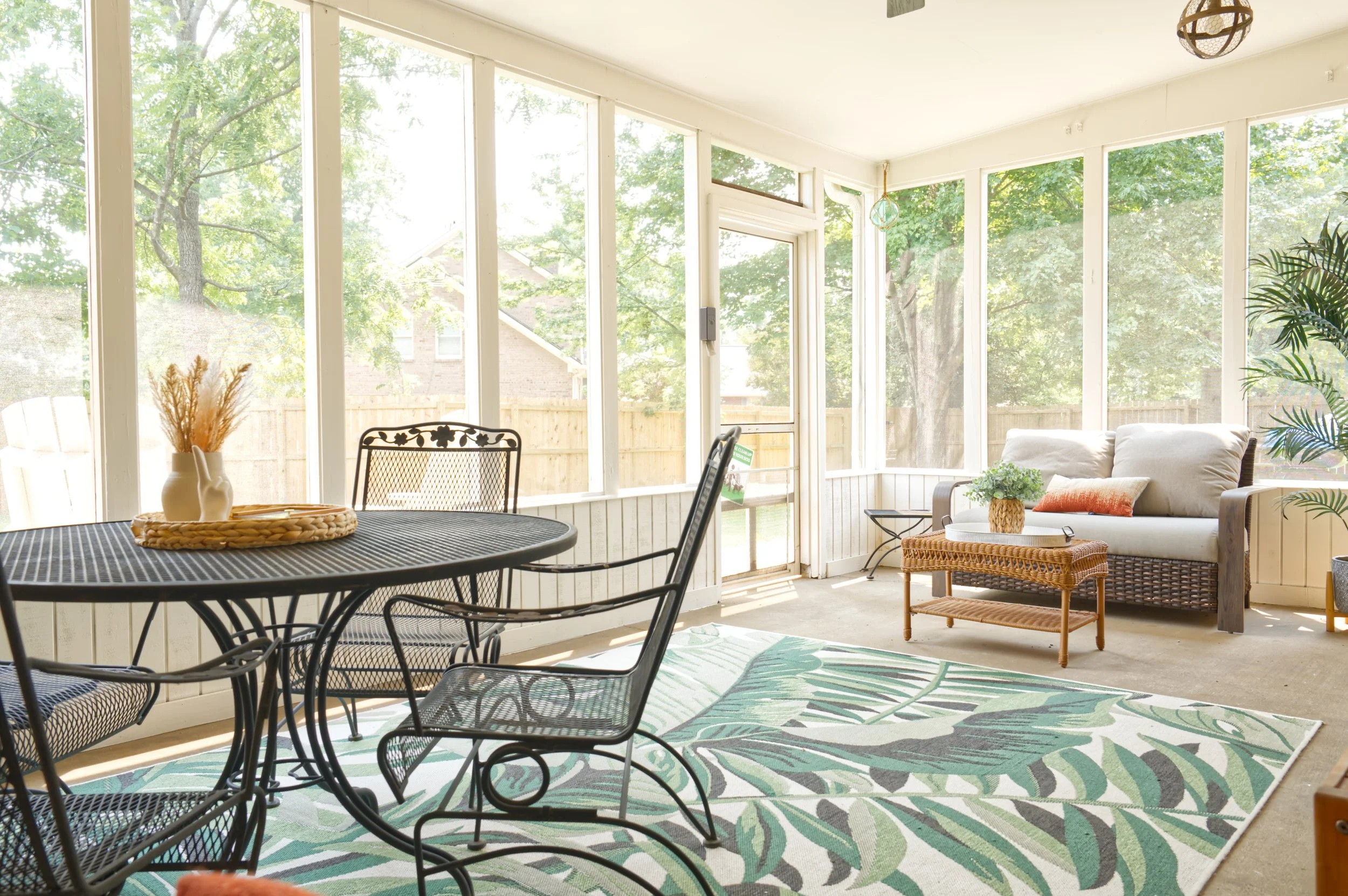 Bright enclosed porch with large windows, a round metal dining table with a woven tray and vases, a white cushioned sofa with pillows, a wicker side table, a patterned outdoor rug, and some greenery including a potted palm.