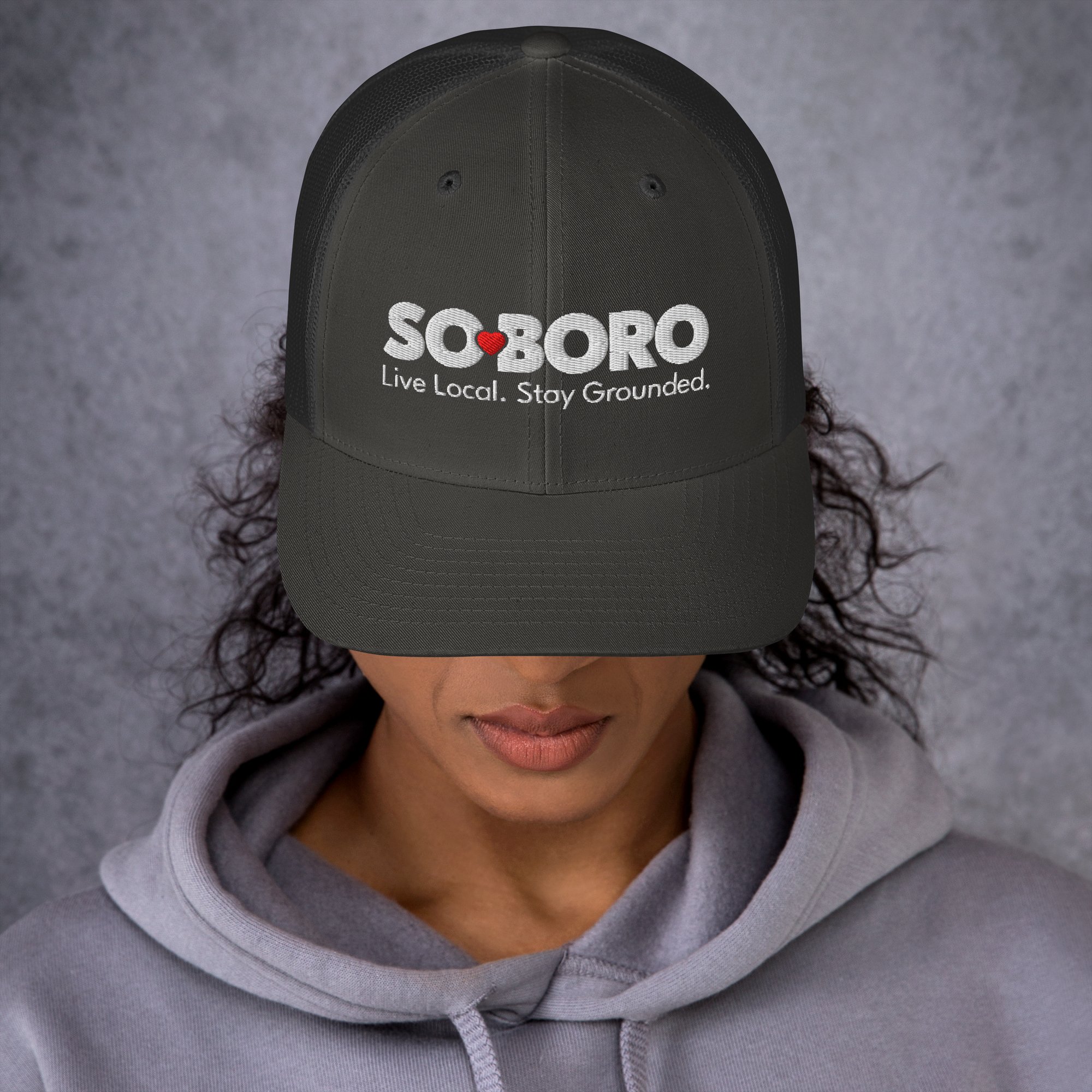 Person wearing a gray hoodie in front of a gray background, with the cap covering their eyes. The cap has the word 'SOBORO' embroidered in white letters, with a red heart replacing the 'O', and the slogan 'Live Local. Stay Grounded.' written below.