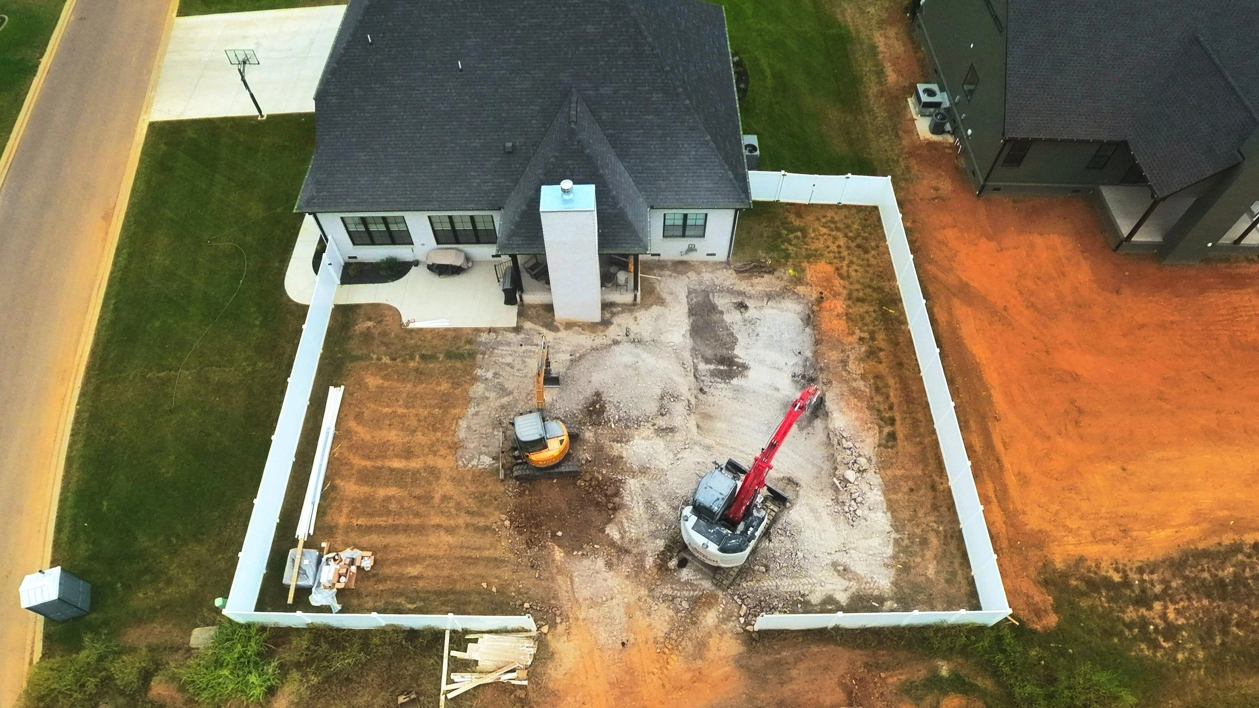 An aerial view of a backyard under construction featuring a house with a black roof, a small patio with outdoor furniture, a fenced yard, and construction equipment including a mini excavator and red crane in the excavated area.