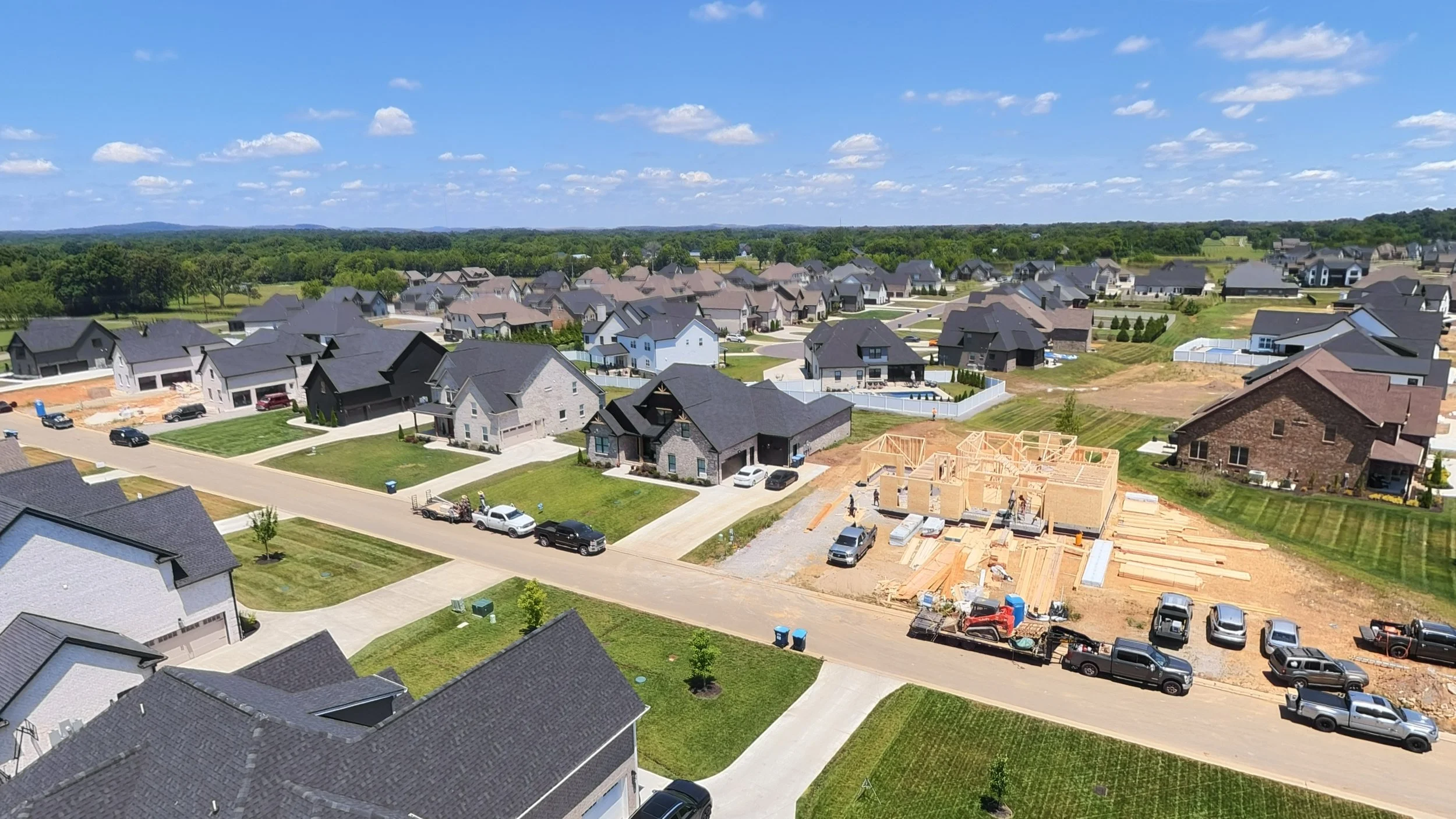 Aerial view of a suburban residential neighborhood with houses, some under construction, and cars parked along the streets on a sunny day with a blue sky and scattered clouds.