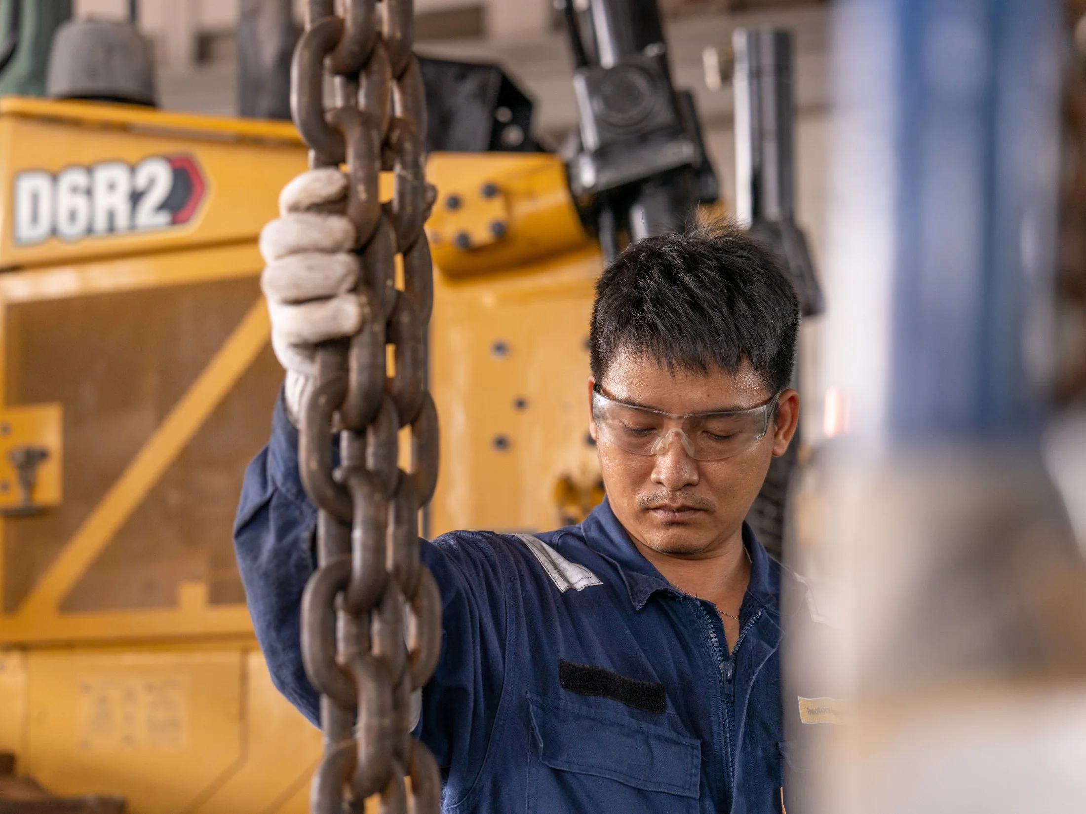 A worker wearing safety glasses and a blue uniform holding a metal chain with a background of industrial equipment and yellow machinery.