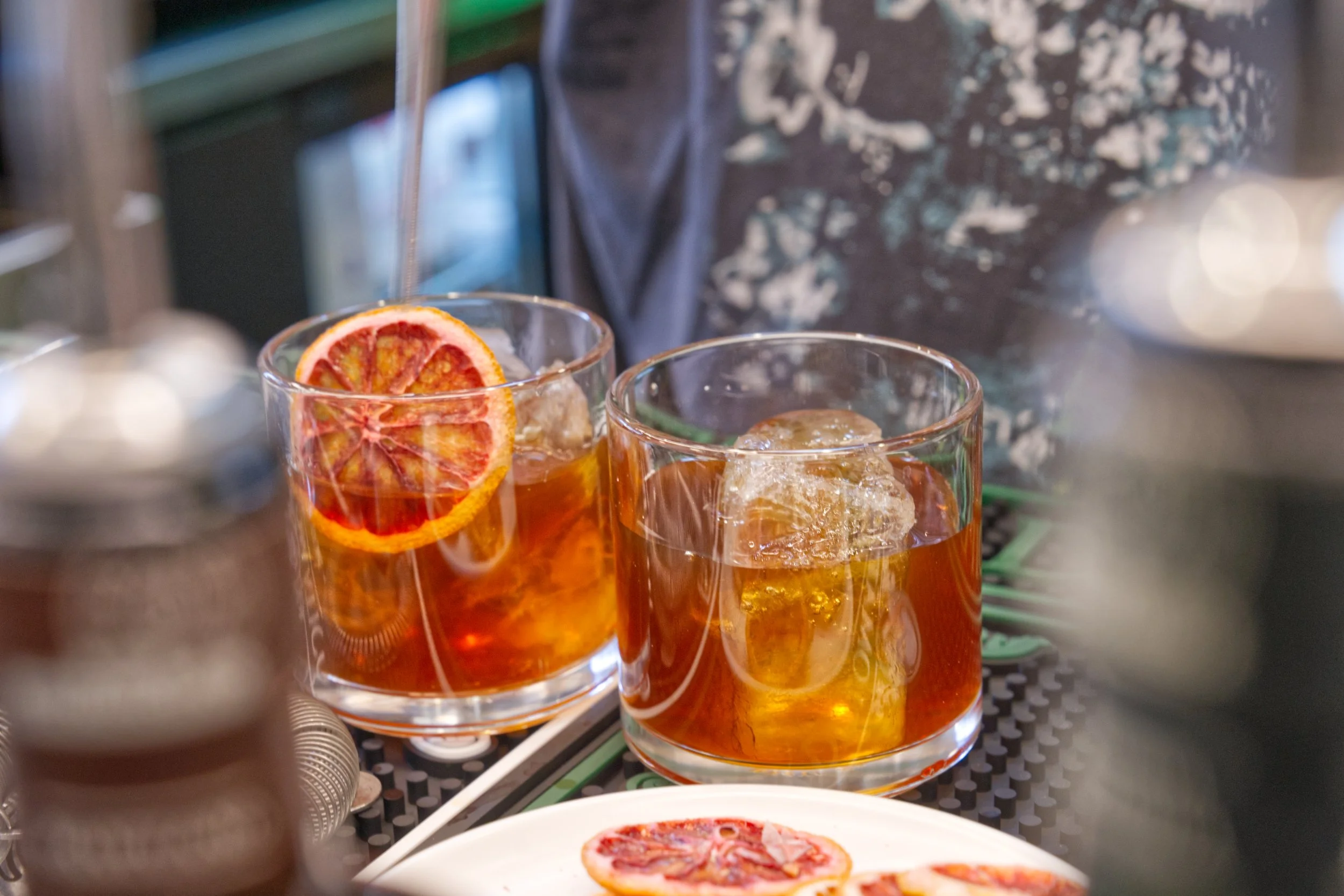 Two glasses filled with an orange-colored beverage and ice, garnished with orange slices, placed on a table. The background shows a person in a dark shirt with a splattered white pattern.