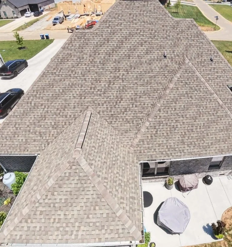 Aerial view of a house with a large shingled roof, neighboring cars, a driveway, a backyard with patio furniture, and a construction site in the background.