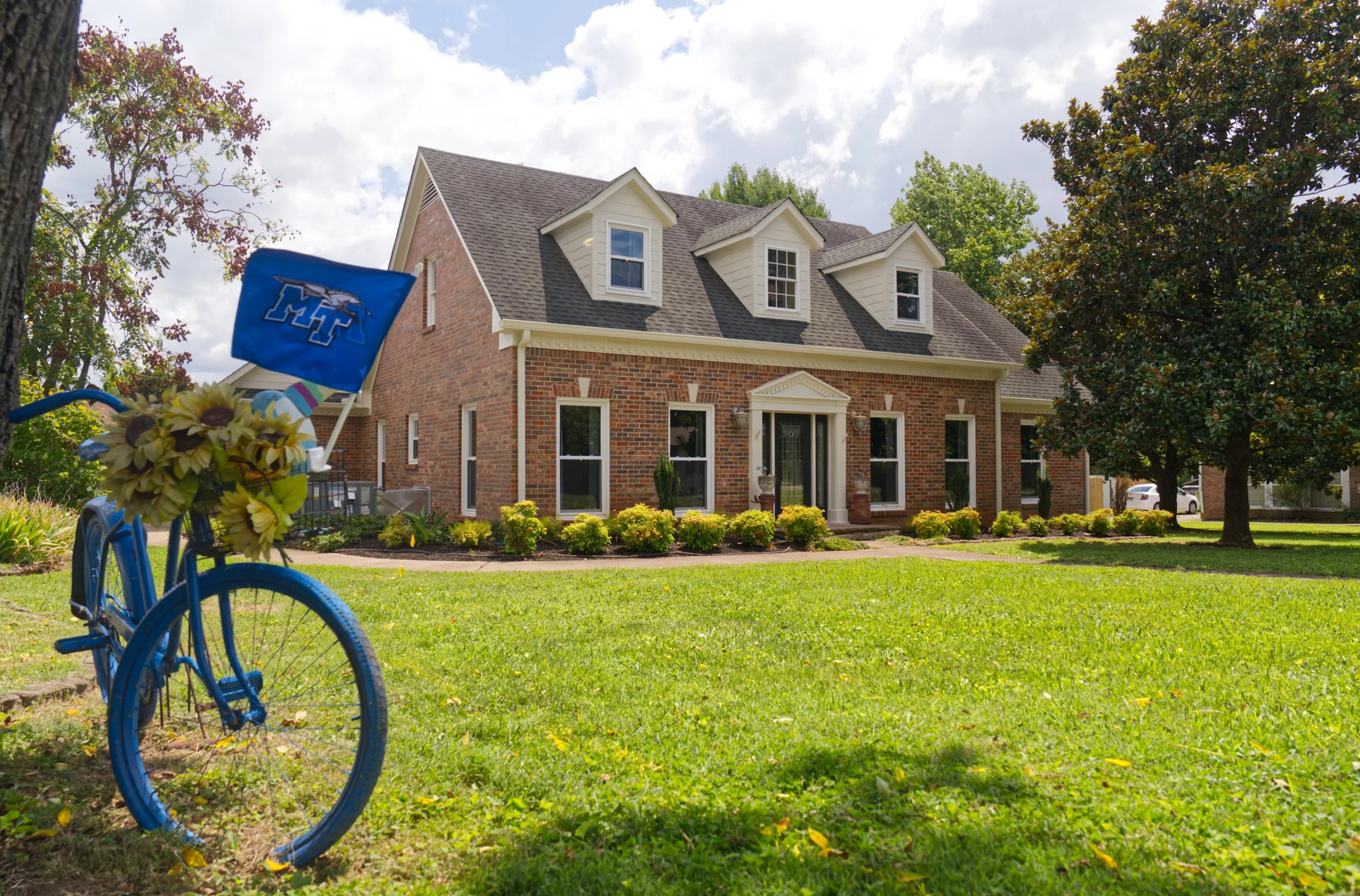 A two-story brick house with a gray shingle roof, three dormer windows, and a front porch, surrounded by green lawn and trees. A blue bicycle with sunflower and rainbow decorations is in the foreground.