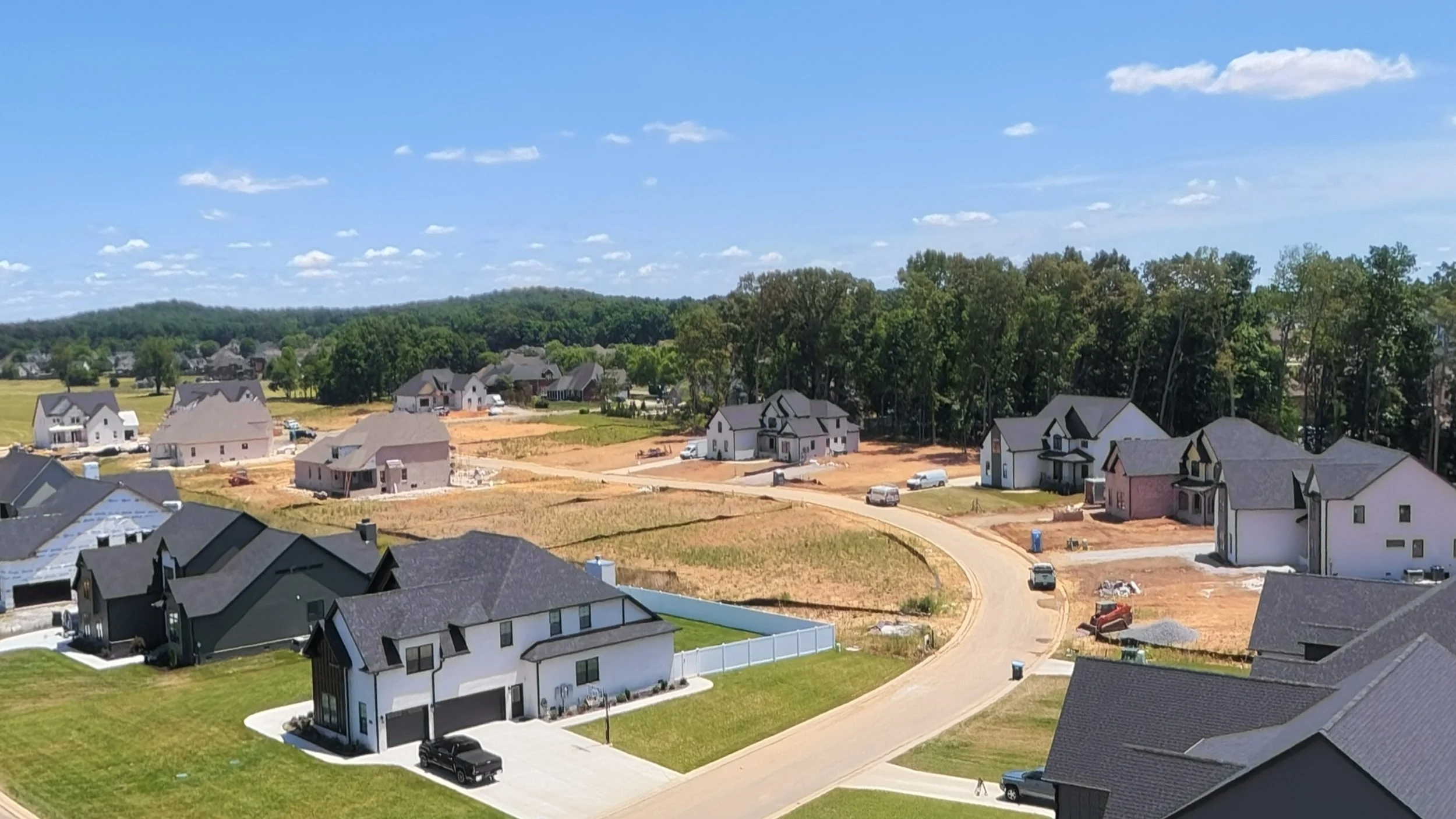 A suburban neighborhood under construction with several houses in various stages of finishing, some with yards and driveways, and a wooded area in the background under a partly cloudy sky.
