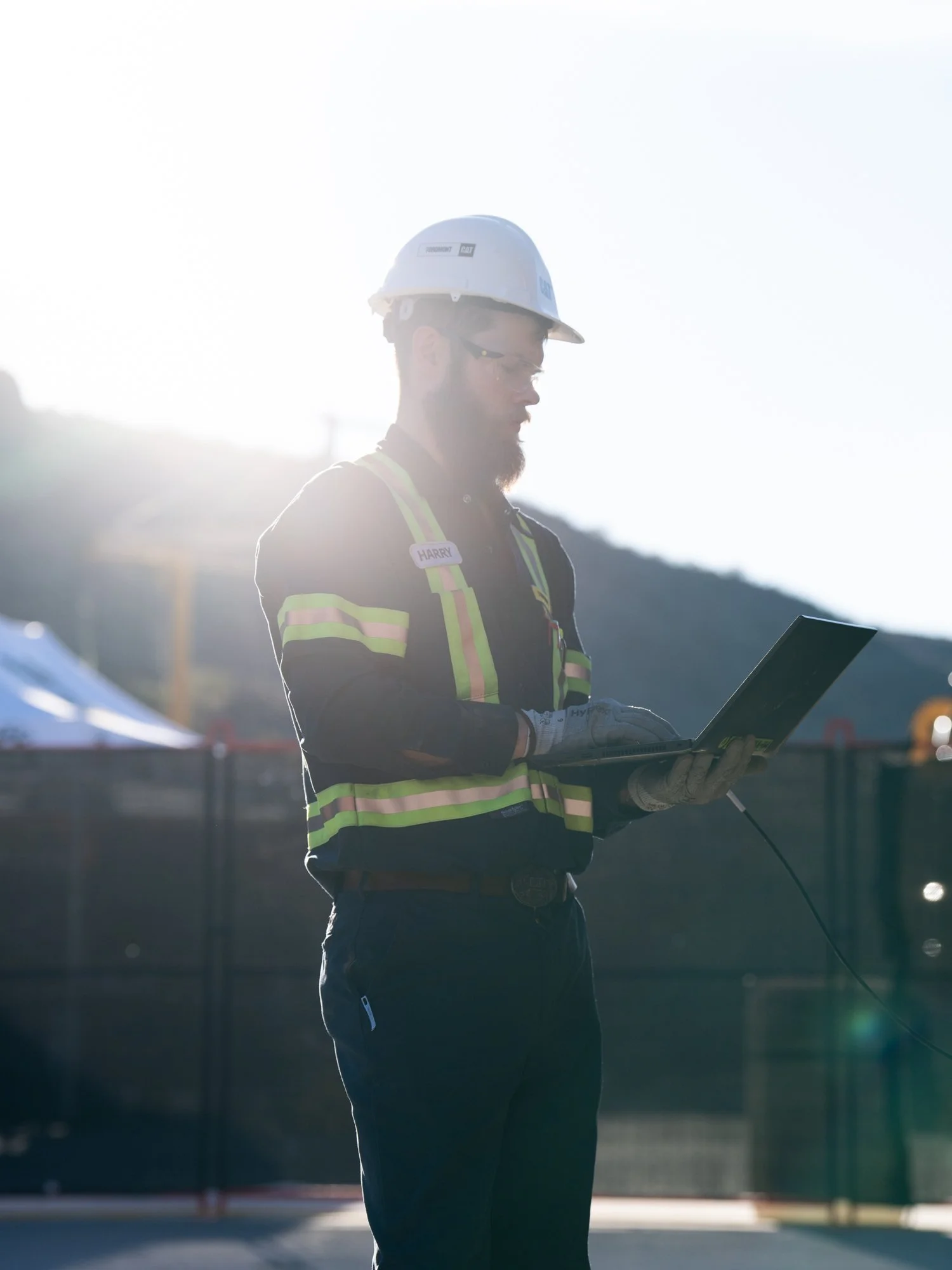 A construction worker wearing a white hard hat and safety vest, standing outdoors and working on a laptop with bright sunlight in the background.