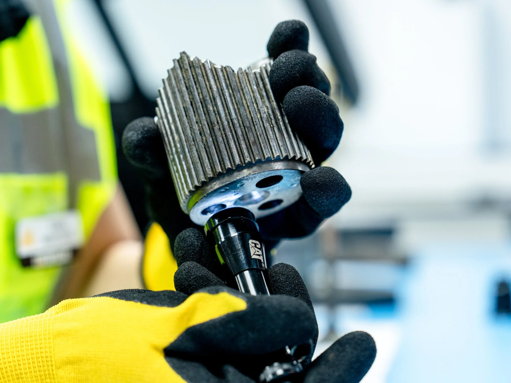 Person wearing yellow gloves and a high-visibility jacket holds a metal gear or sprocket in a workshop setting.