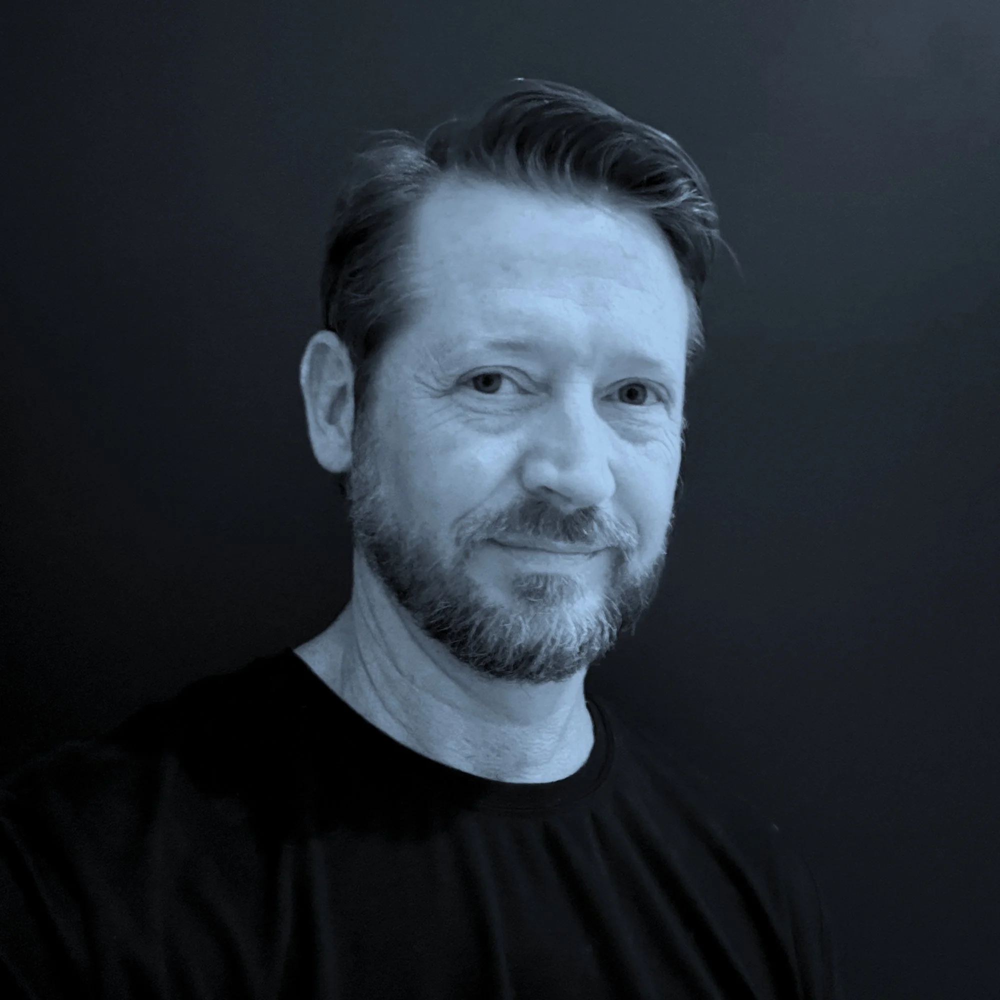Black-and-white photo of a man with styled hair and a beard, looking directly at the camera, with a dark background.