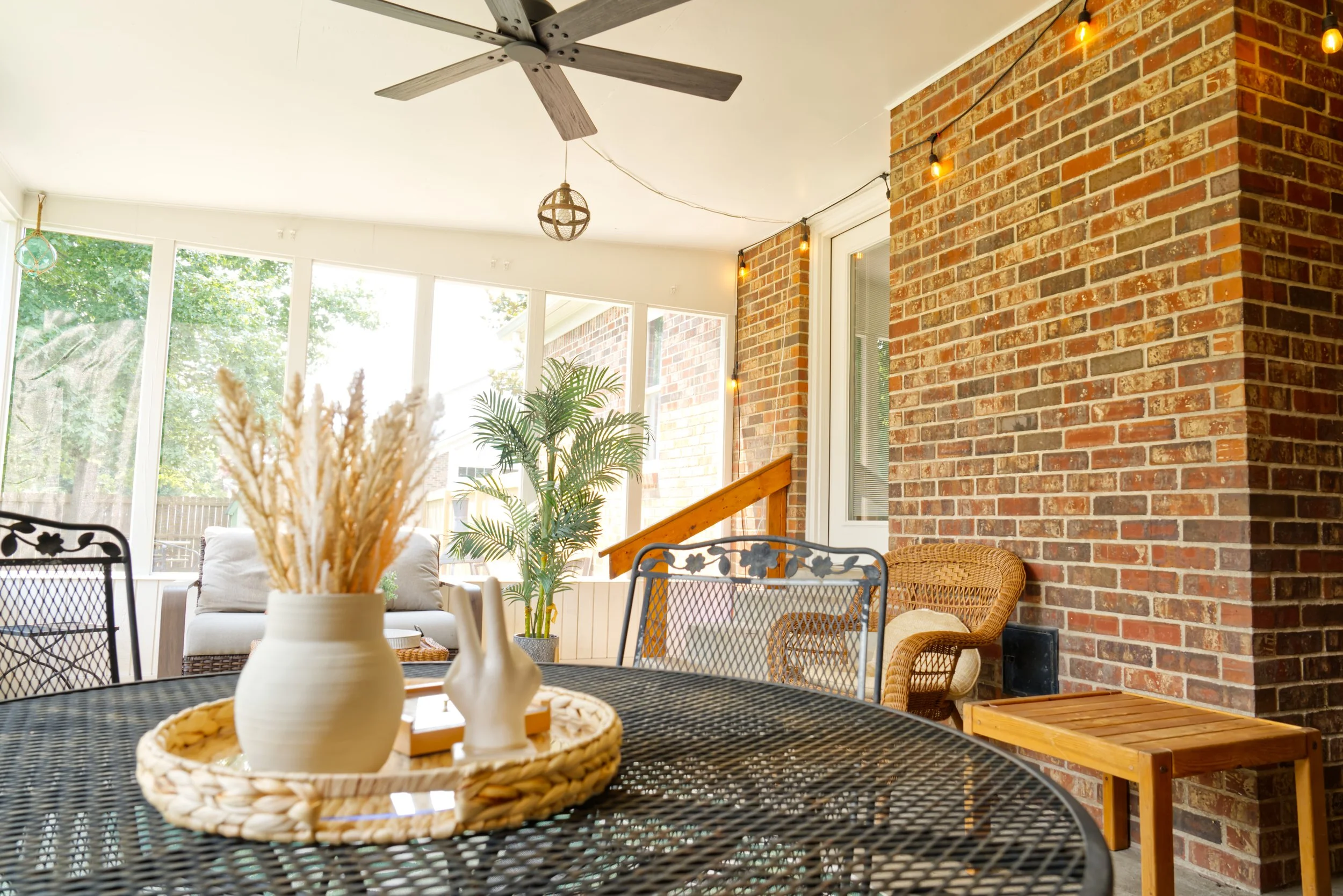 A sunlit porch with wicker furniture, potted plants, a decorative vase, a brick wall with string lights, and a ceiling fan.