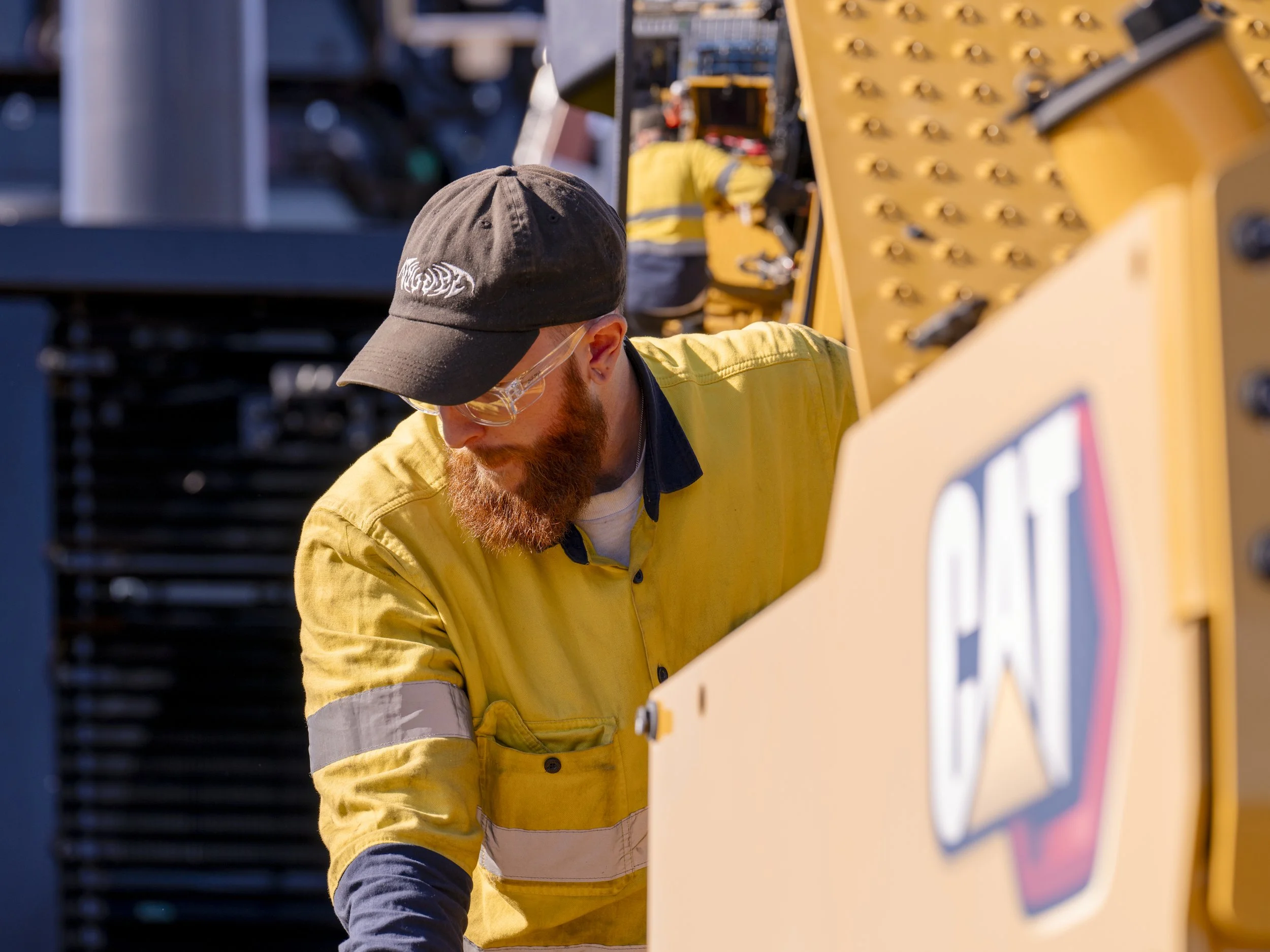 Worker in yellow safety shirt and black cap working on a construction machine.
