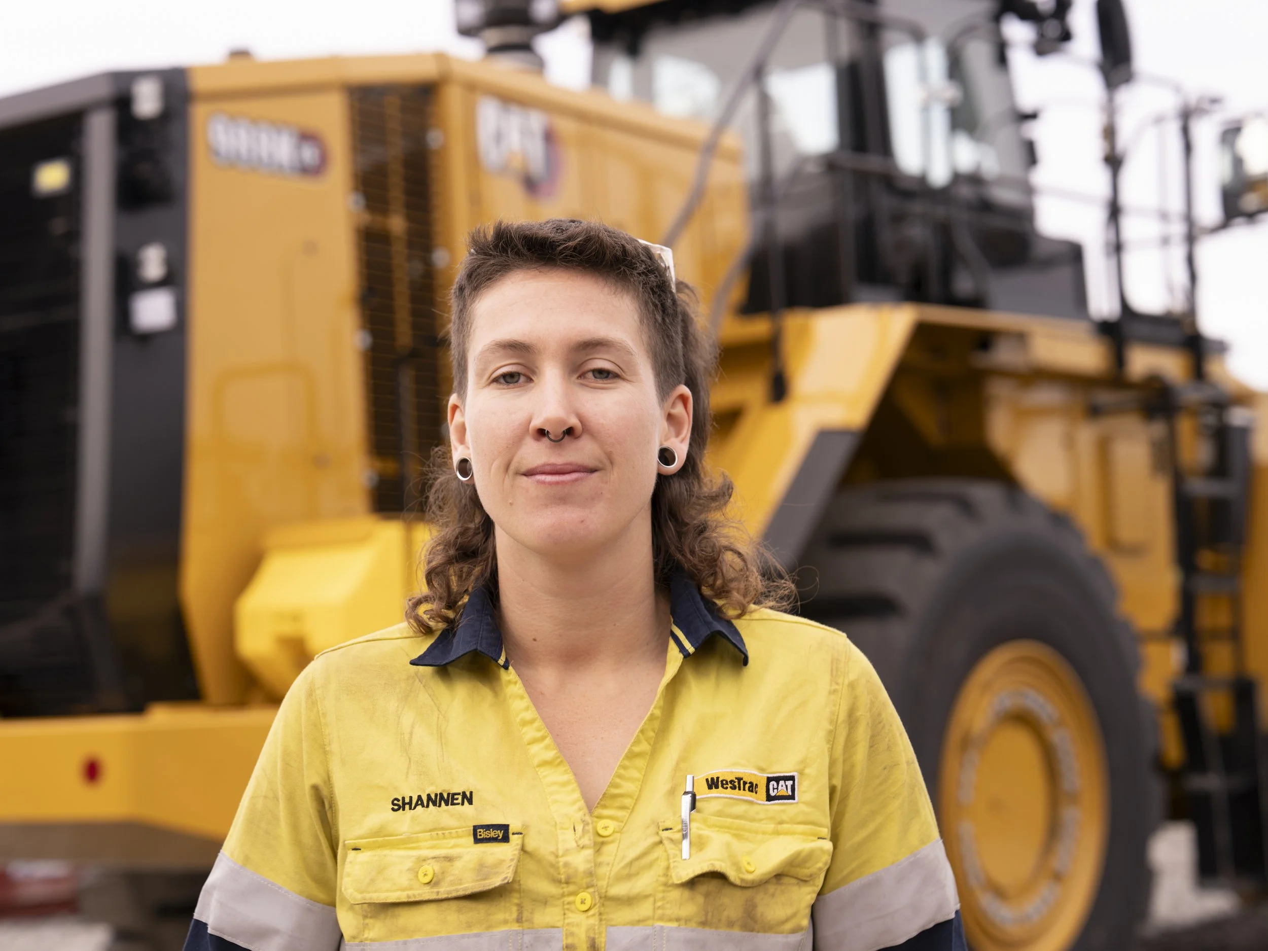 A young woman with curly hair, piercings, and name badges on her yellow work uniform stands in front of large yellow construction equipment.