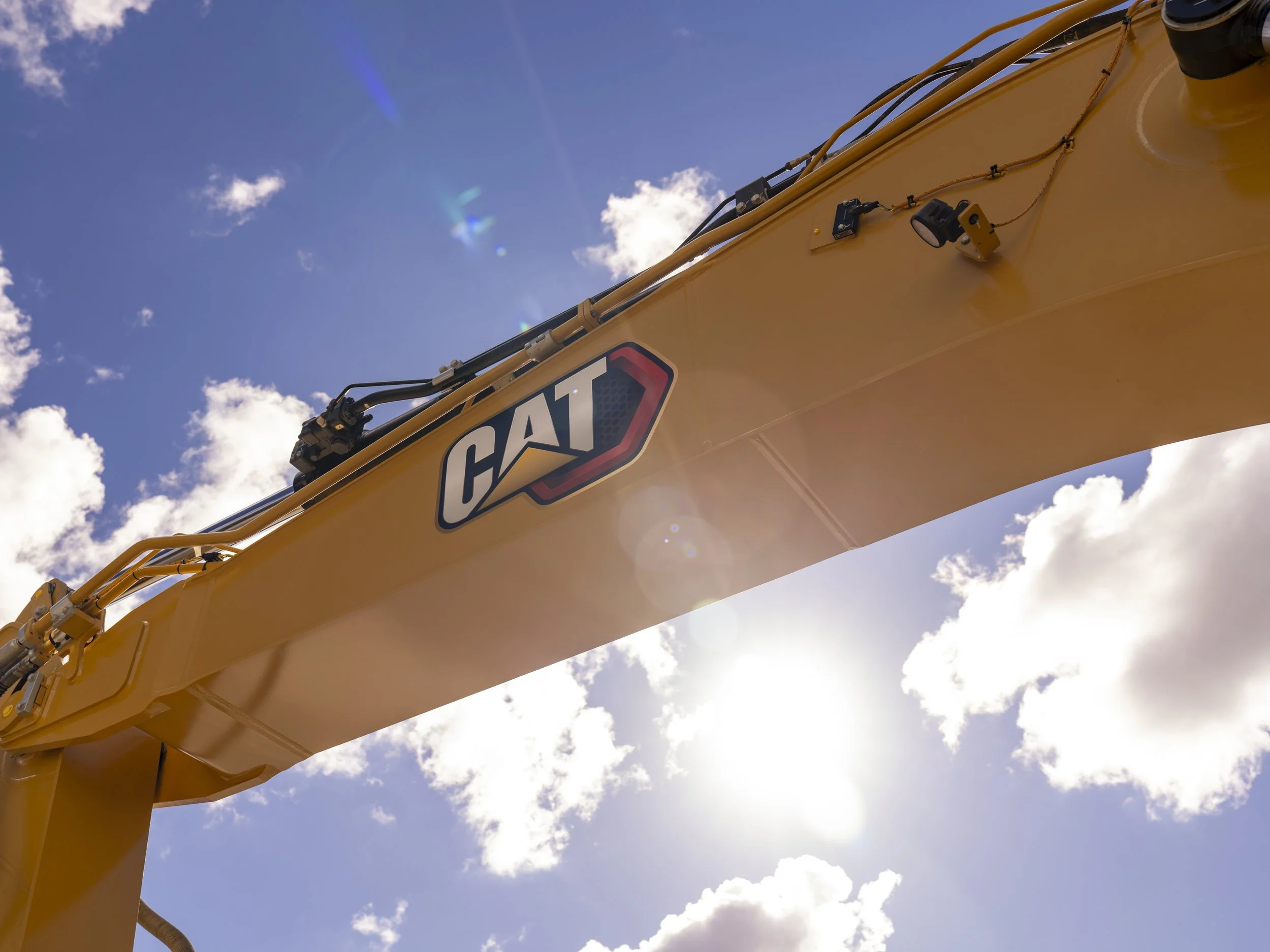 Close-up of a yellow CAT construction excavator arm with hydraulic hoses, against a blue sky with scattered clouds.