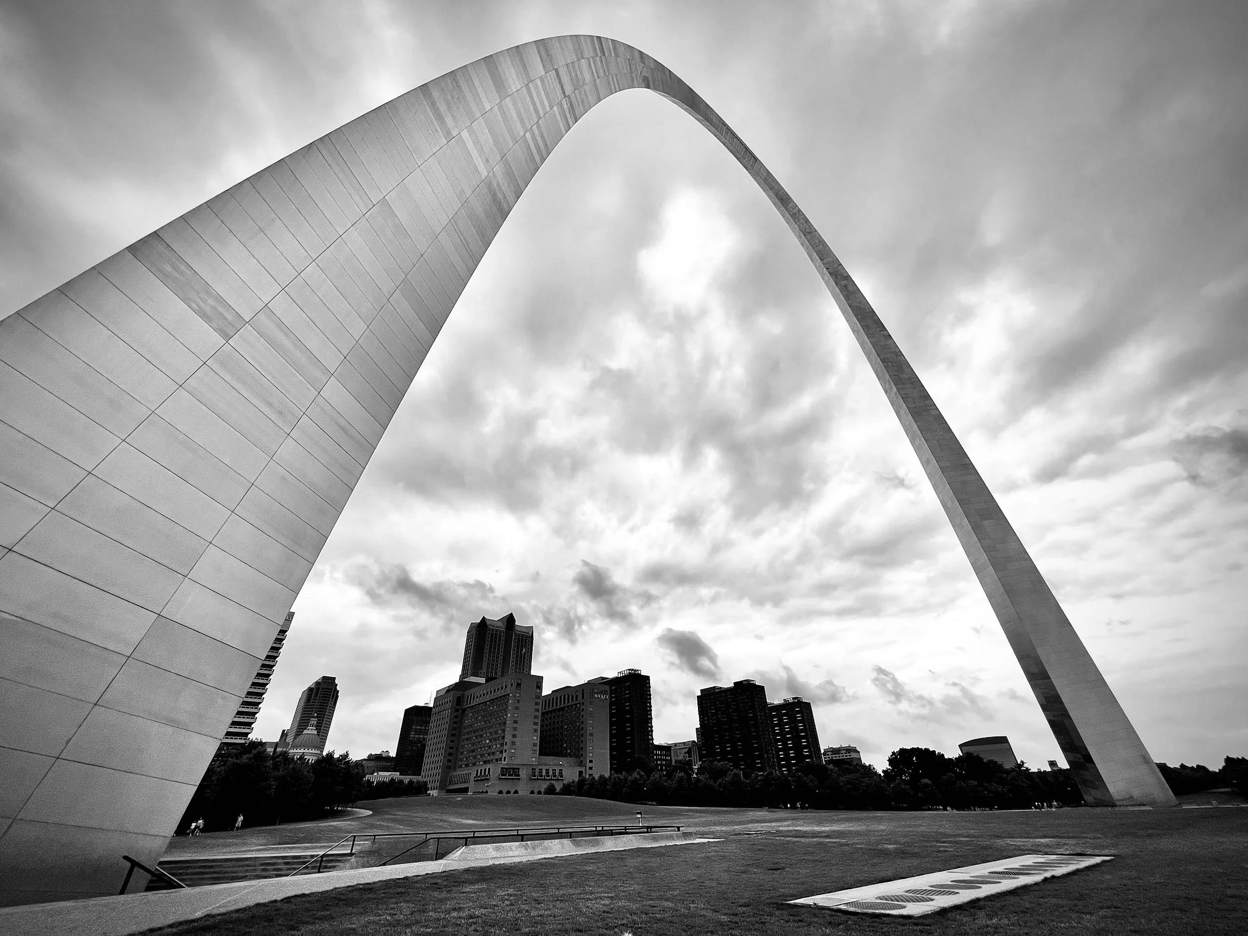 Black and white photo of the Gateway Arch in St. Louis, Missouri, seen from a low angle, with surrounding skyscrapers and park in the foreground under a cloudy sky.