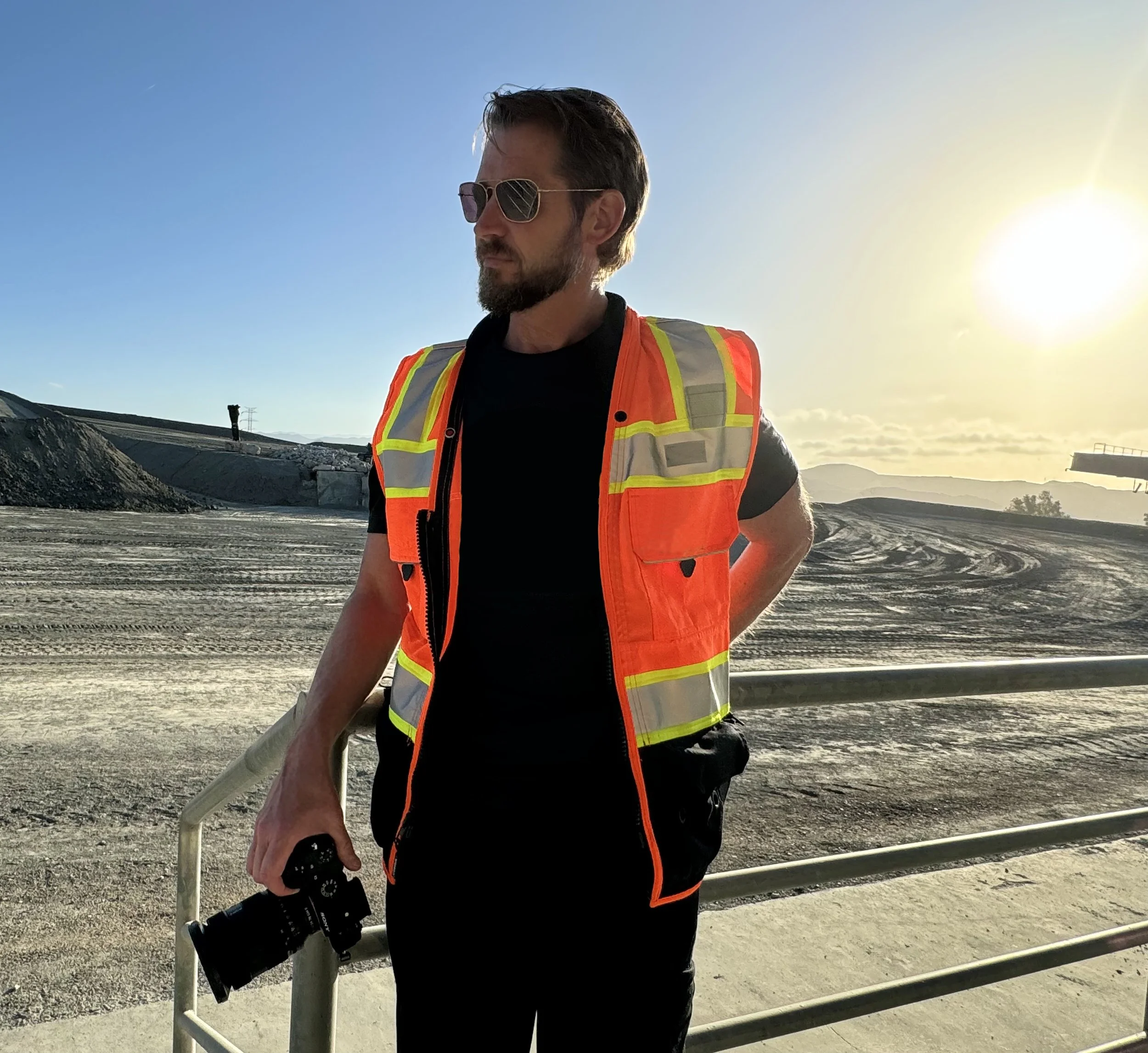 A man wearing sunglasses and an orange safety vest is standing outdoors in a desert or construction site with mountains in the background, holding a camera lens in his right hand, with the sun setting or rising behind him.