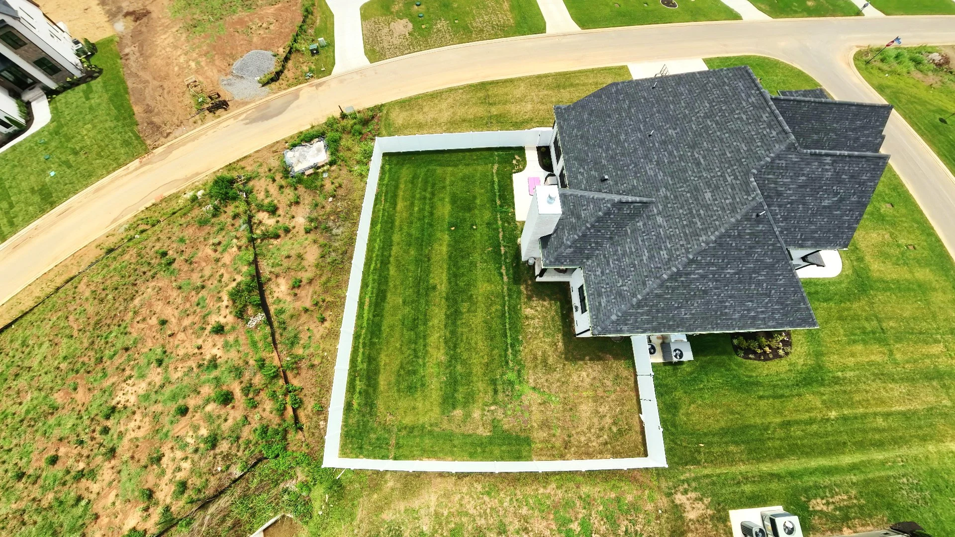 Aerial view of a house with a large backyard, a patio, and a manicured lawn, with neighboring houses and a dirt road surrounding the property.