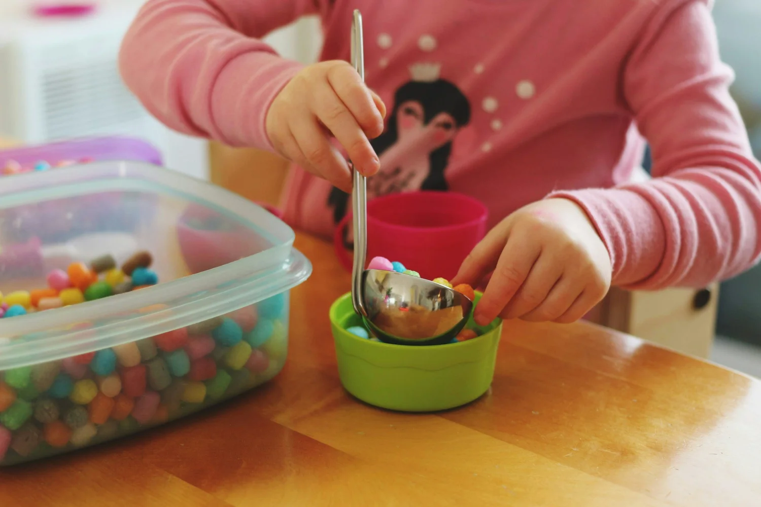 A toddler scooping pom-poms from a sensory bin with tongs