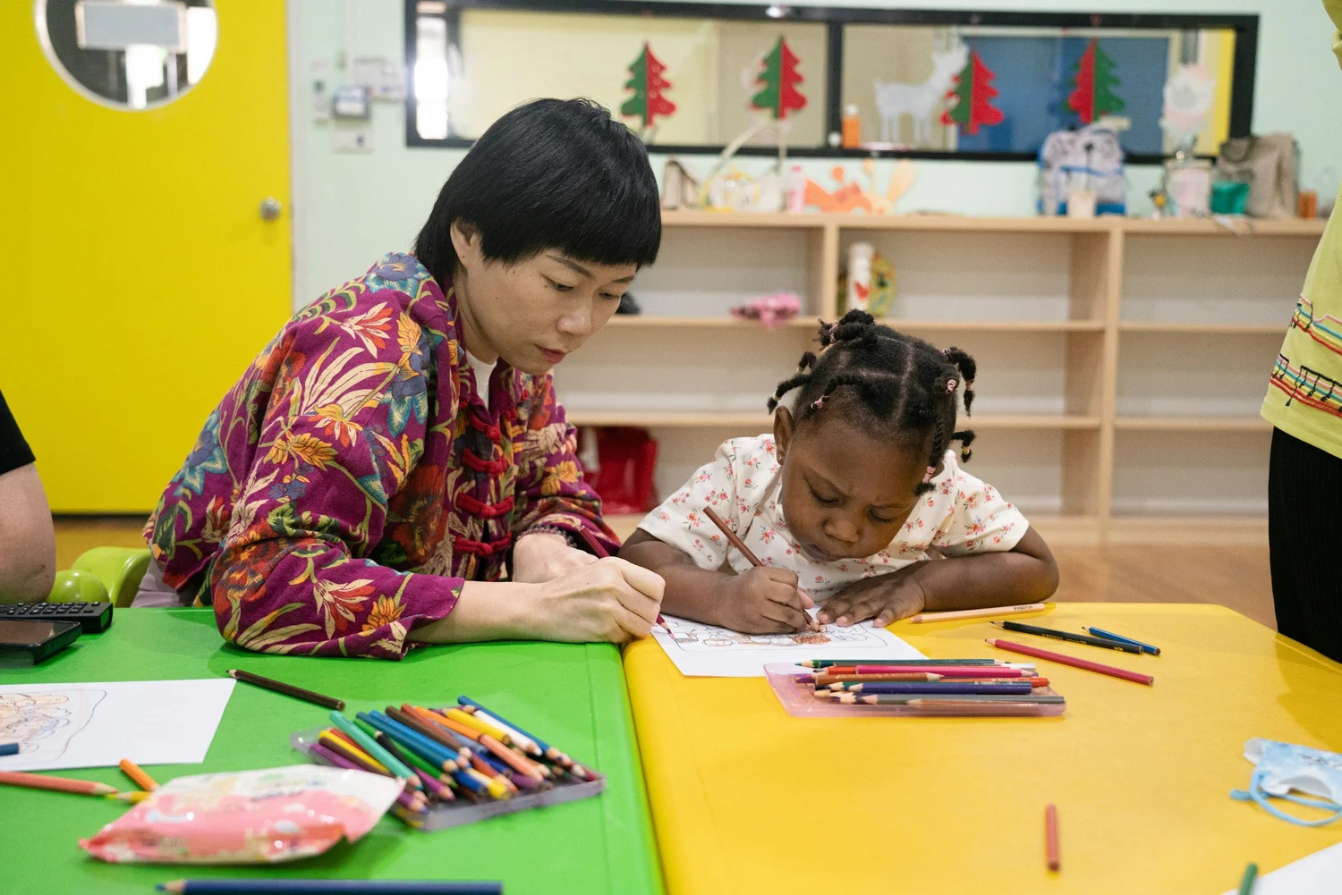 A child practicing writing at a preschool table with a teacher