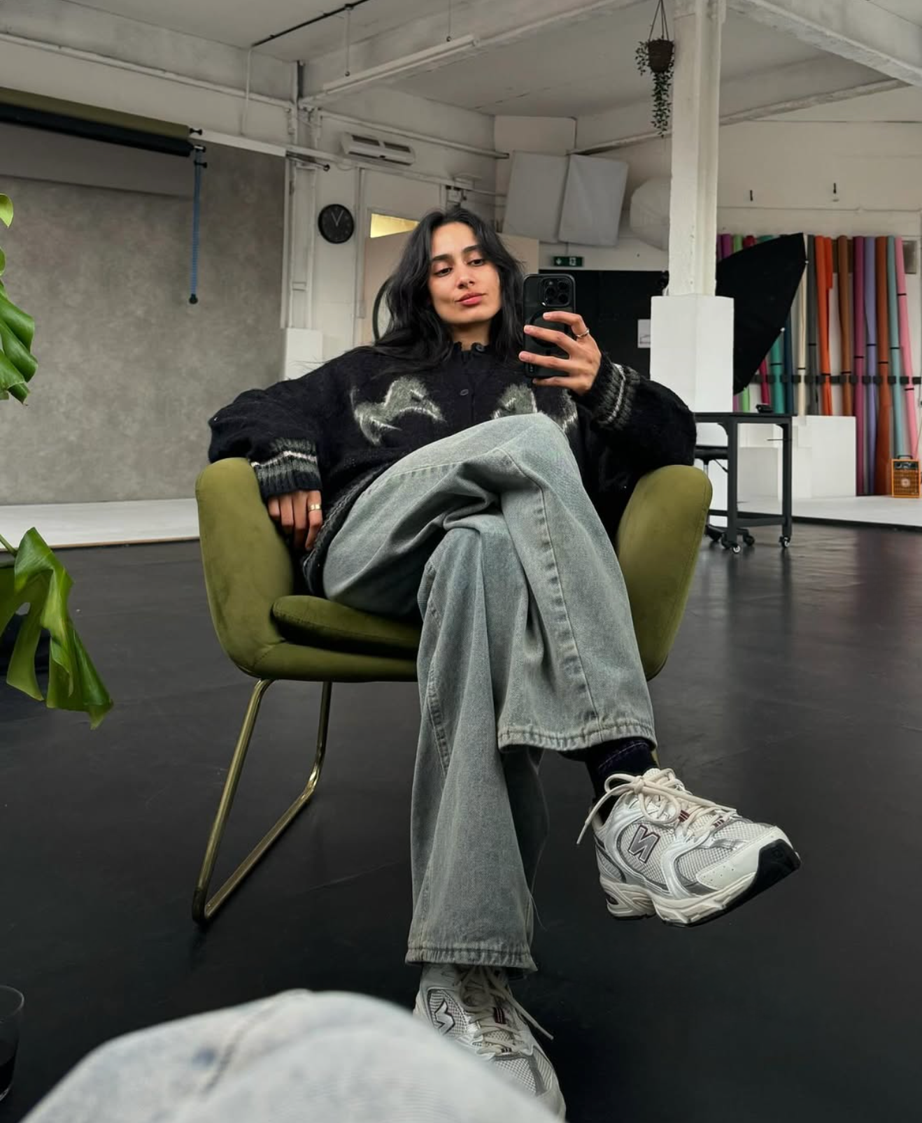 A woman with dark hair sitting in a green chair, taking a selfie in a spacious studio or office with colorful rolled papers on the wall, a black softbox light, a small table with wheels, and a potted plant hanging above.