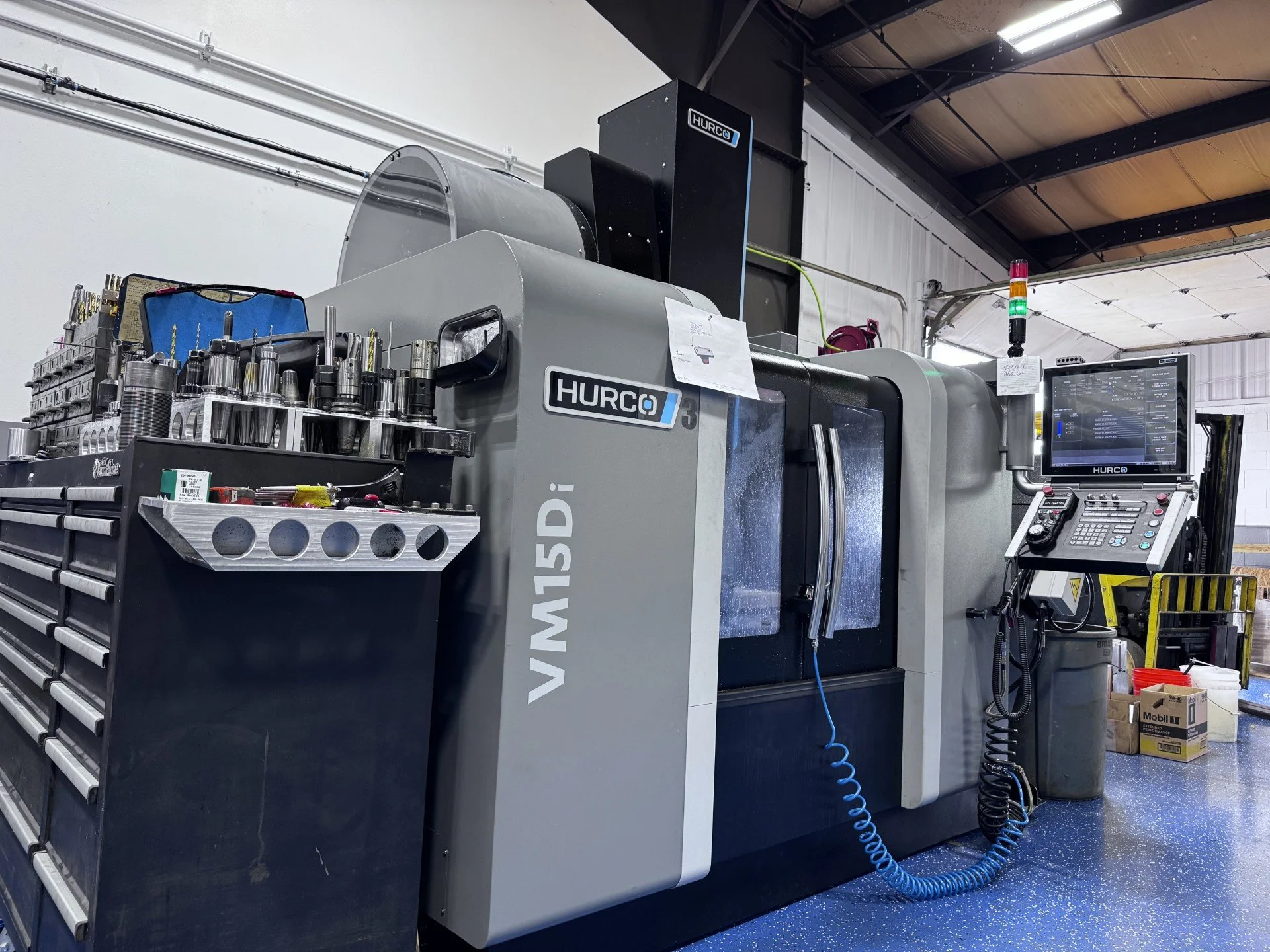 A large industrial metal CNC machine labeled 'VAM-V1015D1' in a factory setting with various tools and equipment around it, including tool holders, a computer monitor, and safety signs.