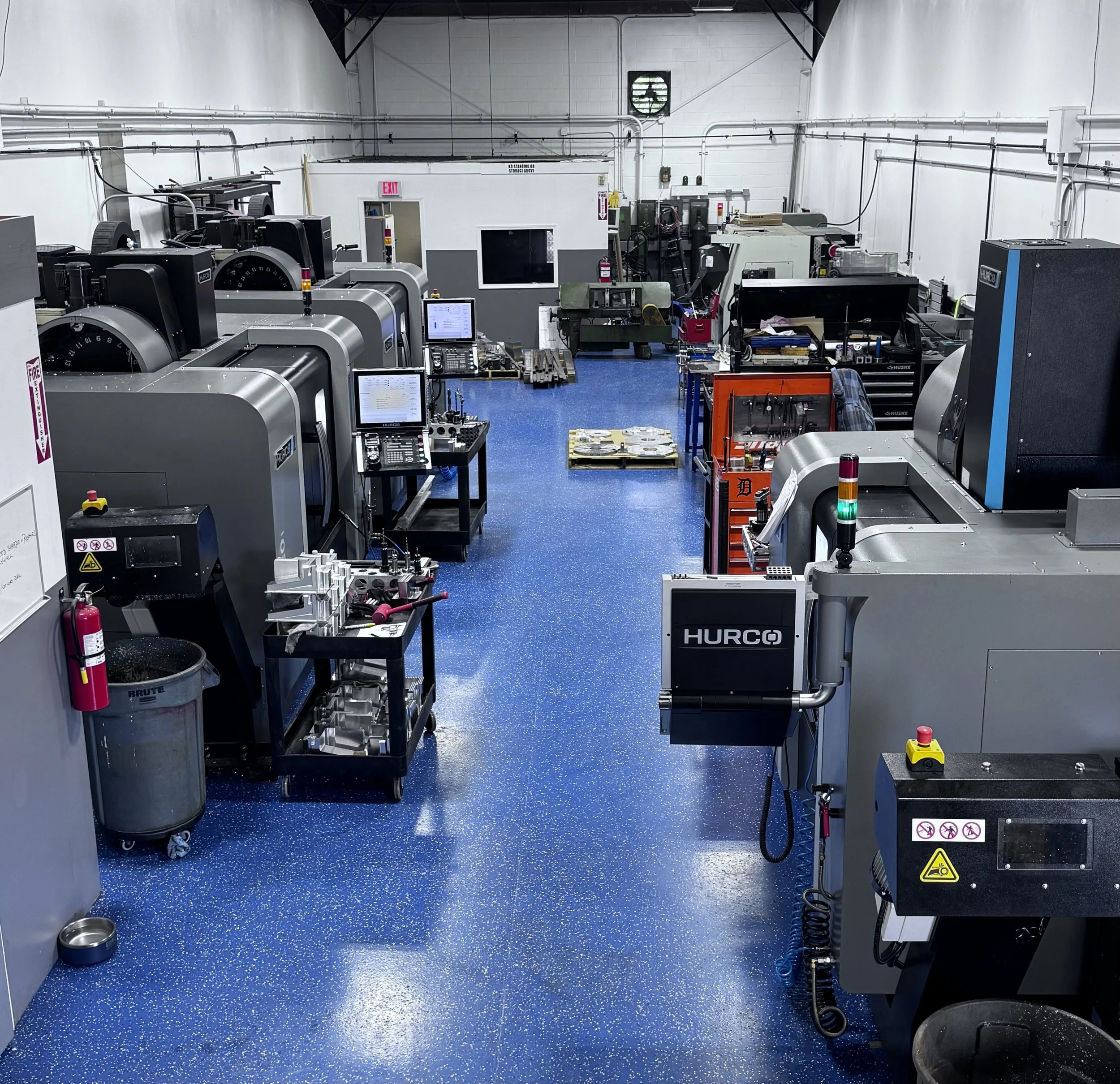 Industrial workshop with CNC machines, workbenches, tools, and equipment on a blue speckled floor and white walls.