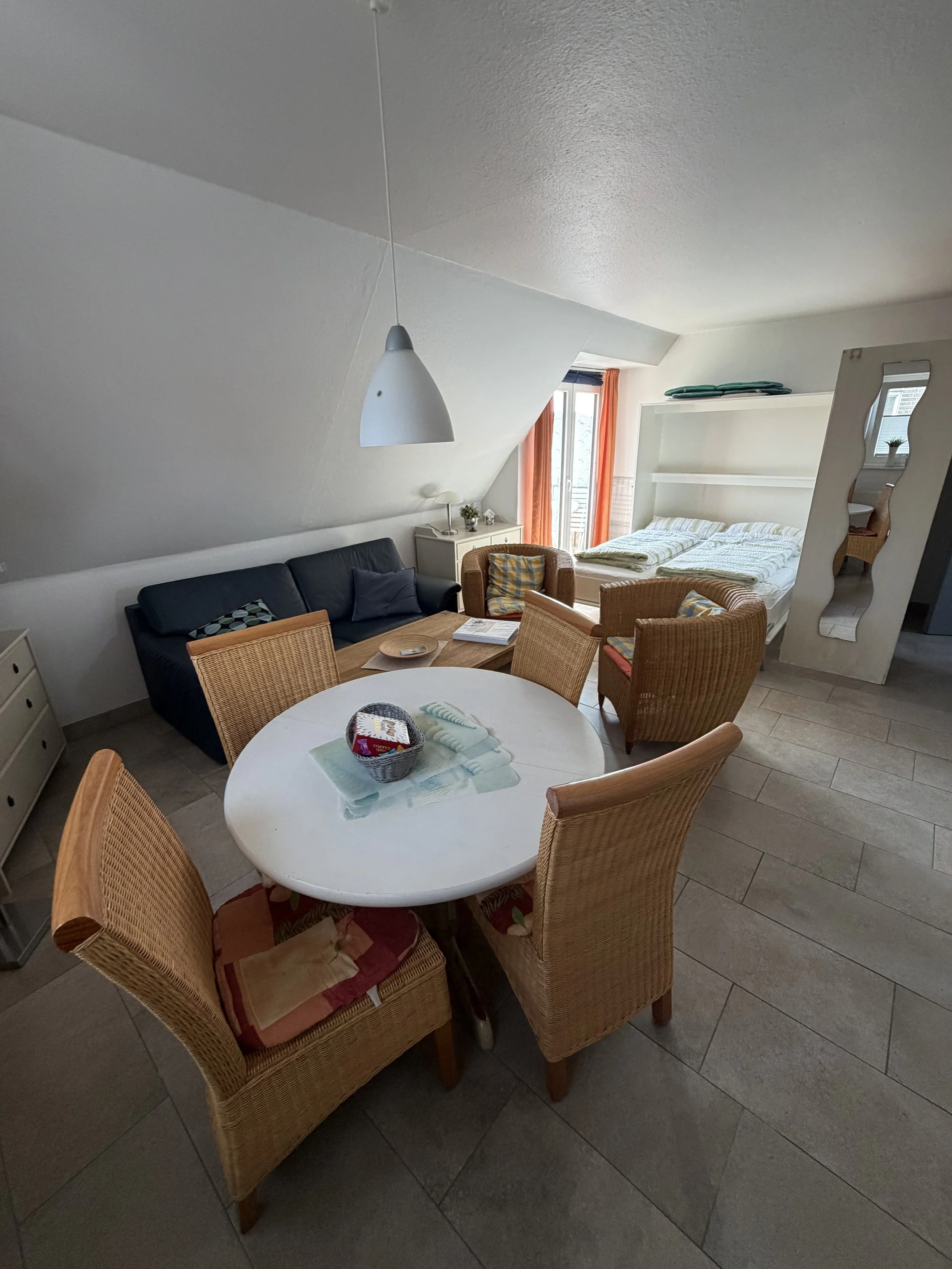 Ein Wohnzimmer mit einem weißen runden Esstisch, sechs Stühlen aus Holzgeflecht, einem Sofa, einem Bett am Fenster, Vorhängen und einem Spiegel.