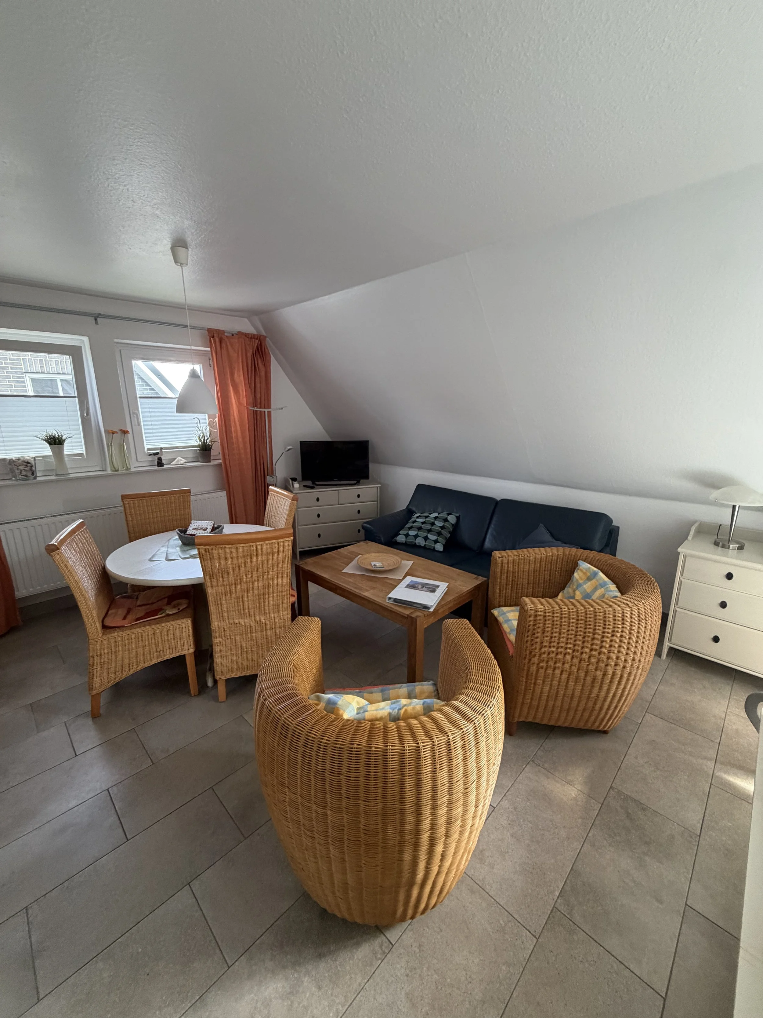 Ein Wohnzimmer mit einem runden Esstisch, sechs Stühlen, einem schwarzen Sofa, einem weißen Sideboard mit Fernseher, mehreren Pflanzen und Vorhängen vor den Fenstern.