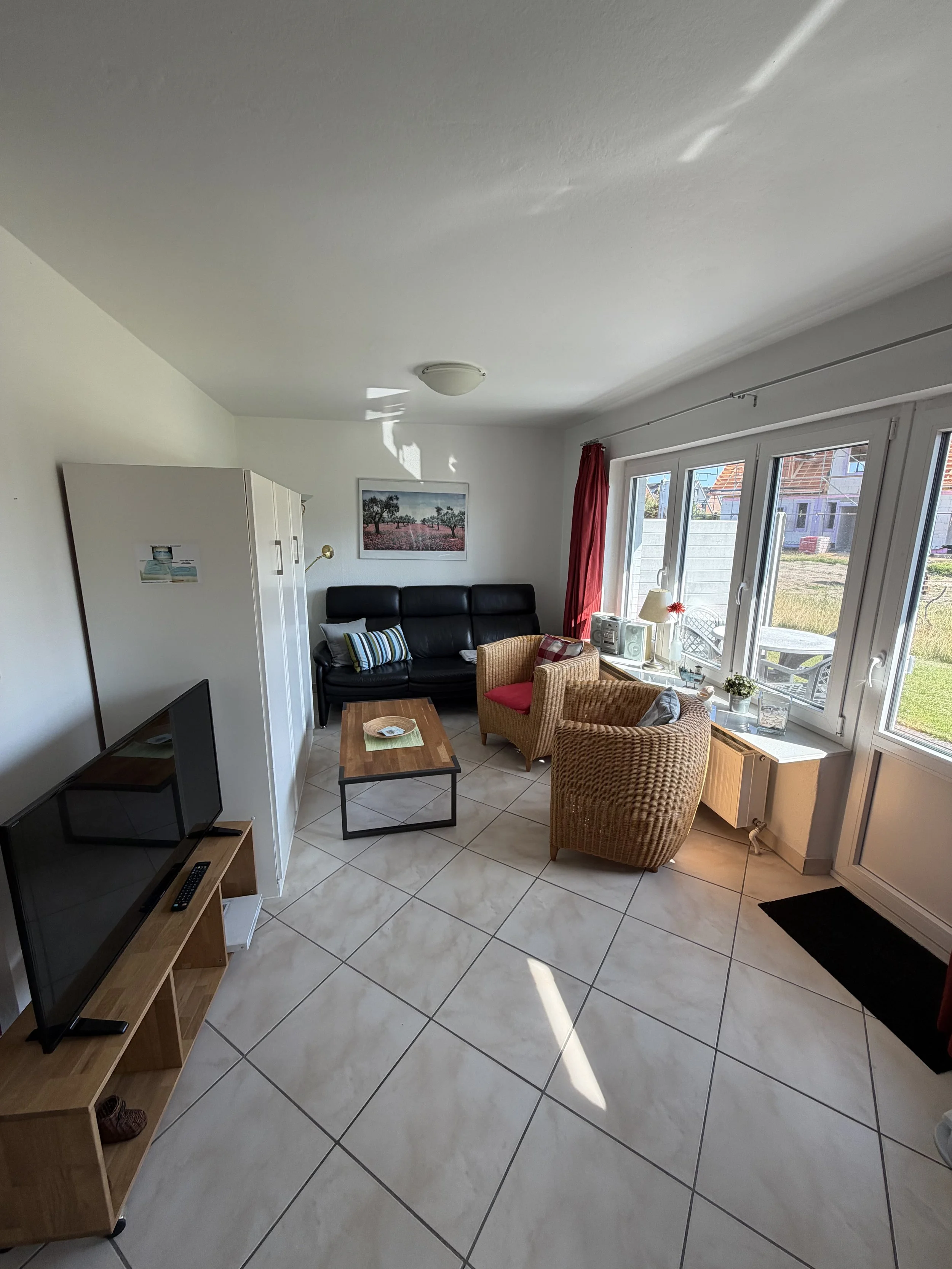 Gemütliches Wohnzimmer mit Sofa, Sessel, TV, Couchtisch, großen Fenstern, Pflanzen und Dekorationen, sonnendurchflutet