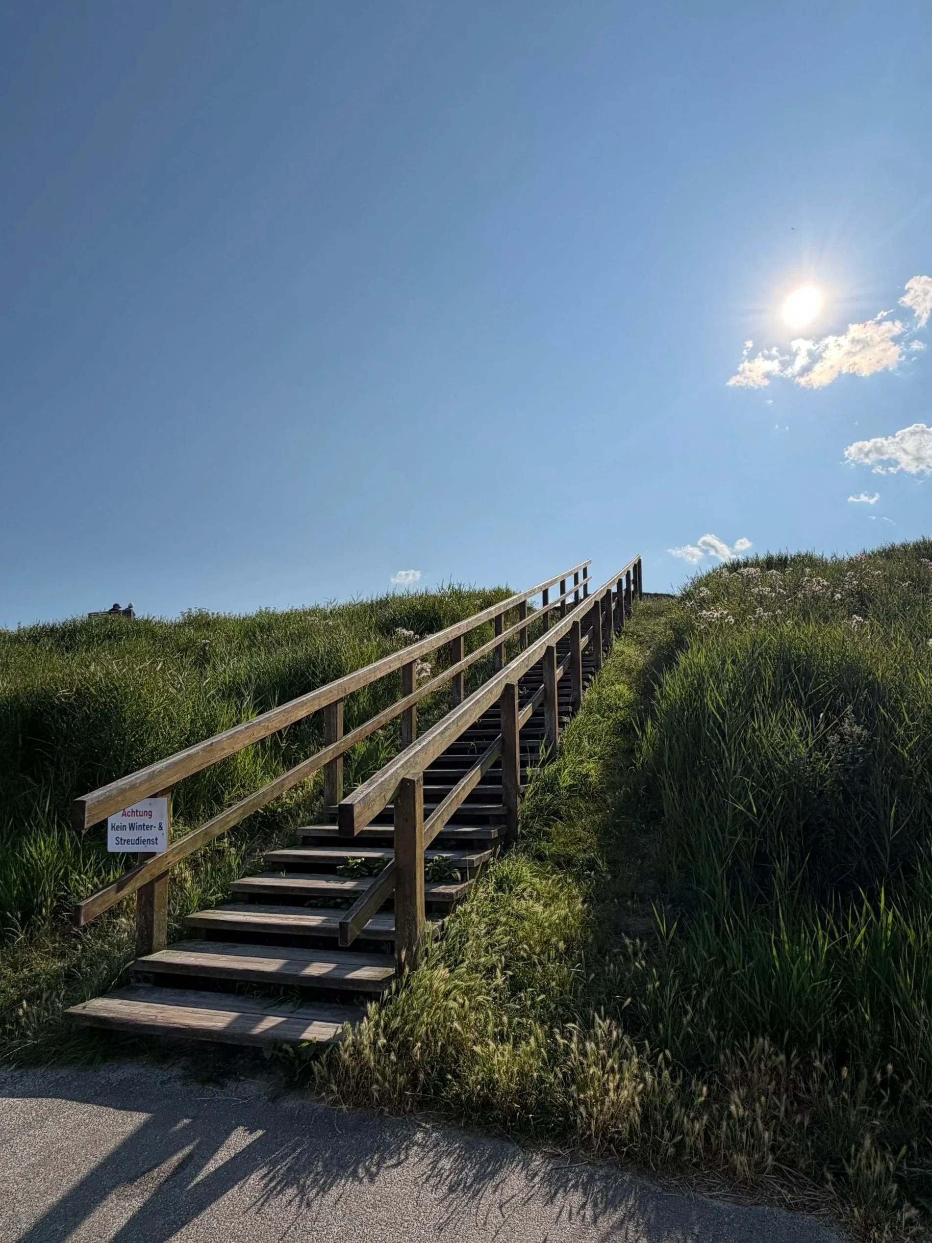 Holztreppe führt durch eine grüne Wiese zum Strand, mit Sonnenlicht am Himmel und einem kleinen Schild, das vor Winter- und Streudienst warnt.