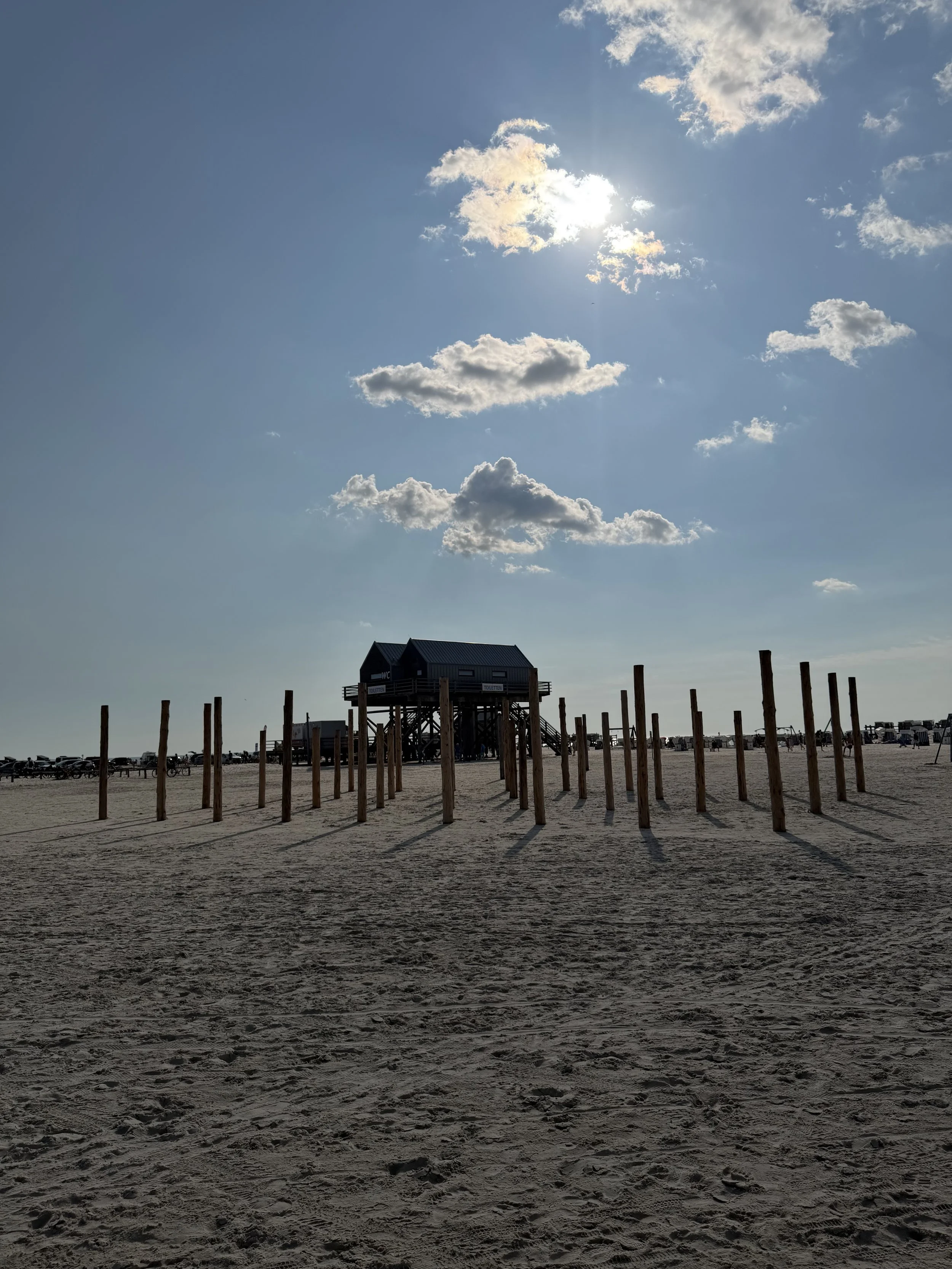 Ein Strand mit einem hohen Holzturm und mehreren Holzpfosten, im Hintergrund sind Sonnenschirme und Liegestühle, der Himmel ist mit einigen Wolken und der Sonne. Es ist wahrscheinlich eine Ferien- oder Strandlandschaft.