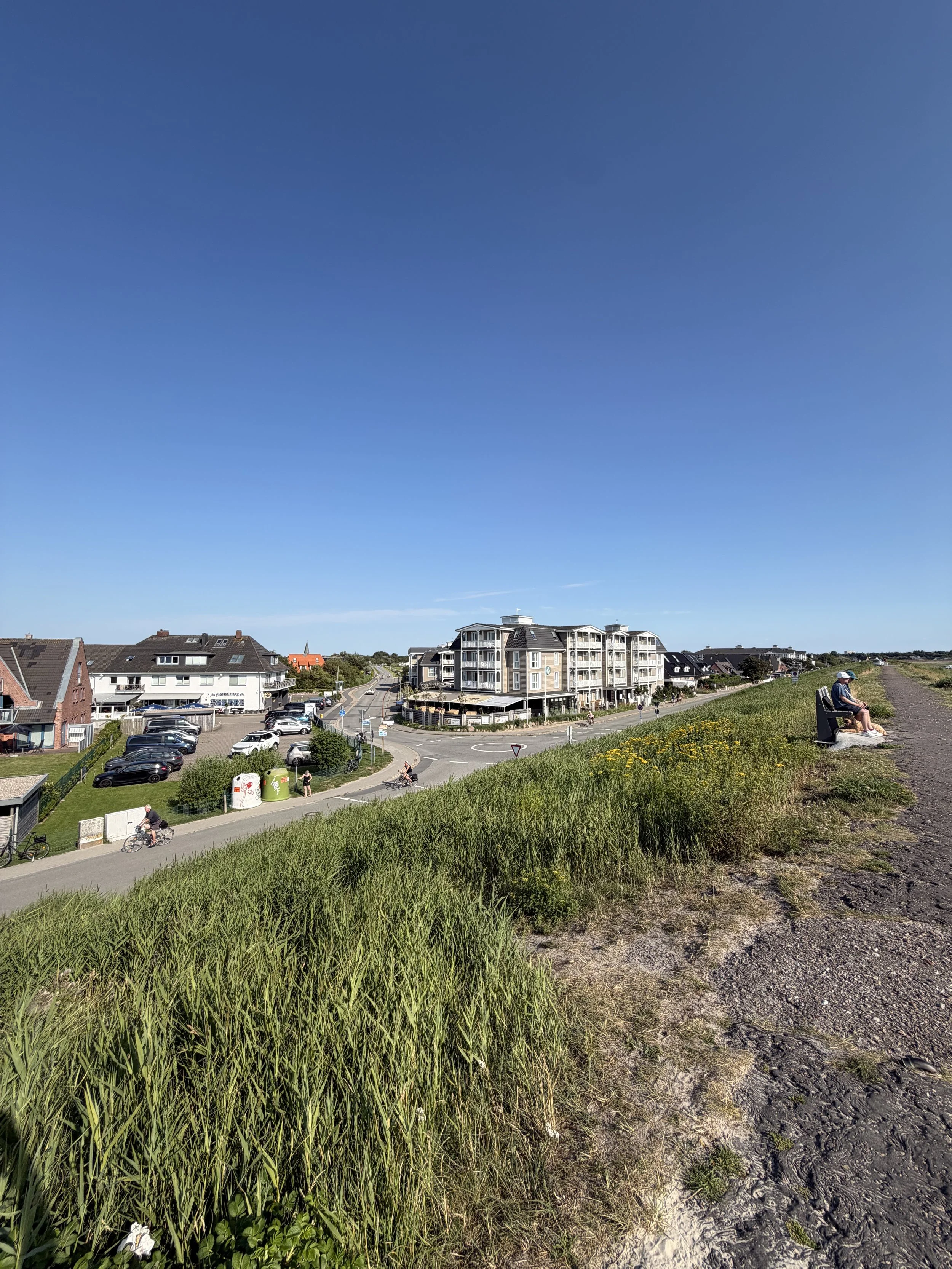 Blick auf eine Wohnanlage mit mehreren Gebäuden, einem Parkplatz und einer Straße. Im Vordergrund ist eine grasbewachsene Böschung mit einem Wanderweg und einer Bank, auf der eine Person sitzt.