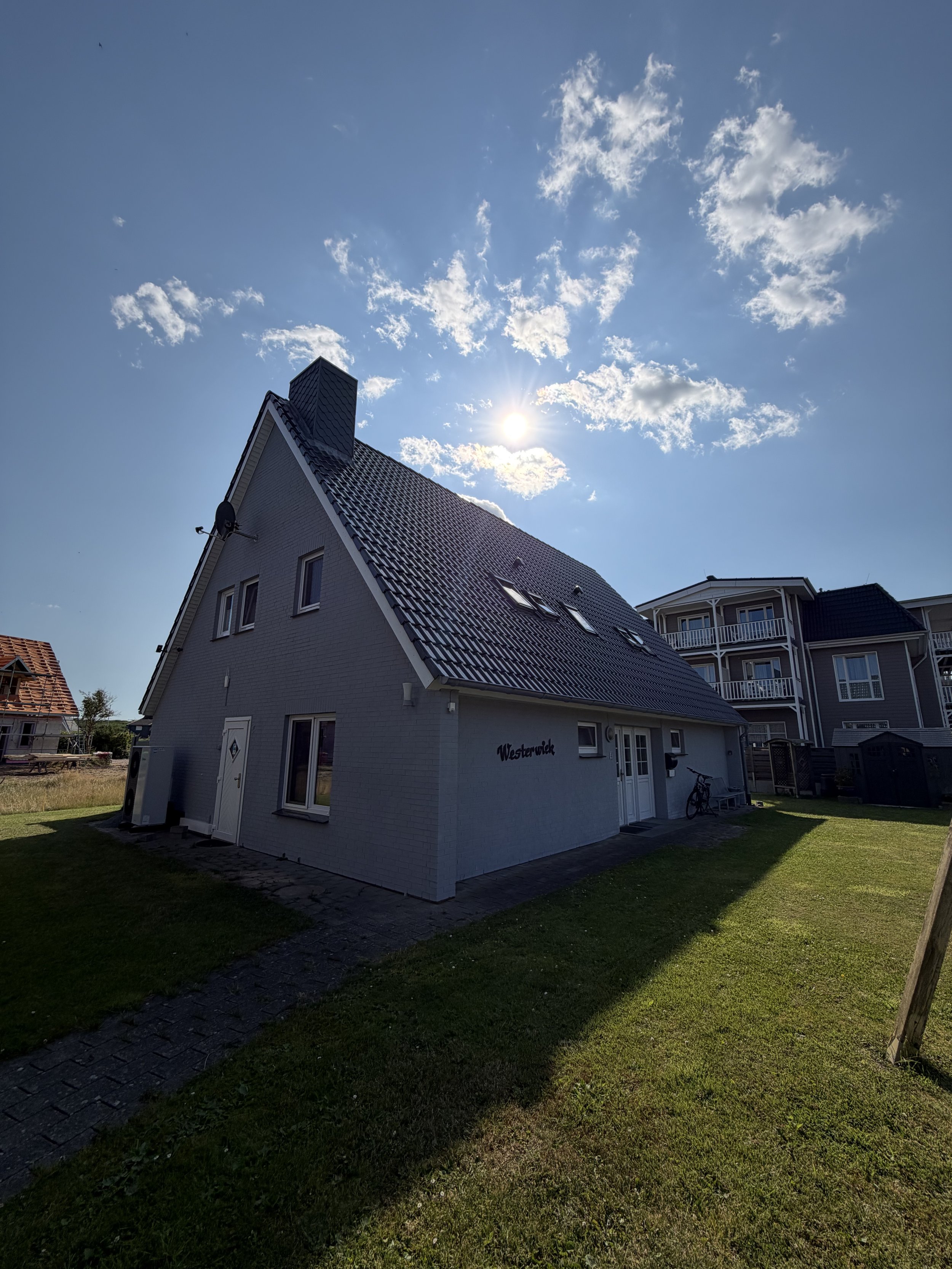 Ein zweistöckiges Haus mit weißen Wänden und dunklem Ziegeldach, im Vordergrund ein grüner Garten mit Schatten, im Hintergrund andere Häuser und ein blauer Himmel mit Wolken und Sonne.