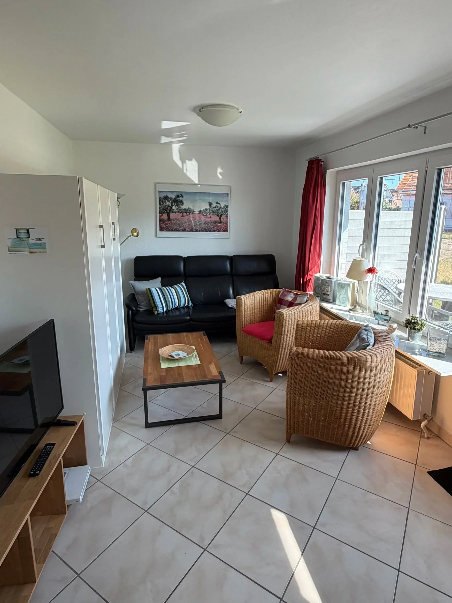 Wohnzimmer mit schwarzen Sofas, Roststuhl, Couchtisch, Kerze, Fenster mit roten Vorhängen, Wandbild, Fernseher und Holzregal.