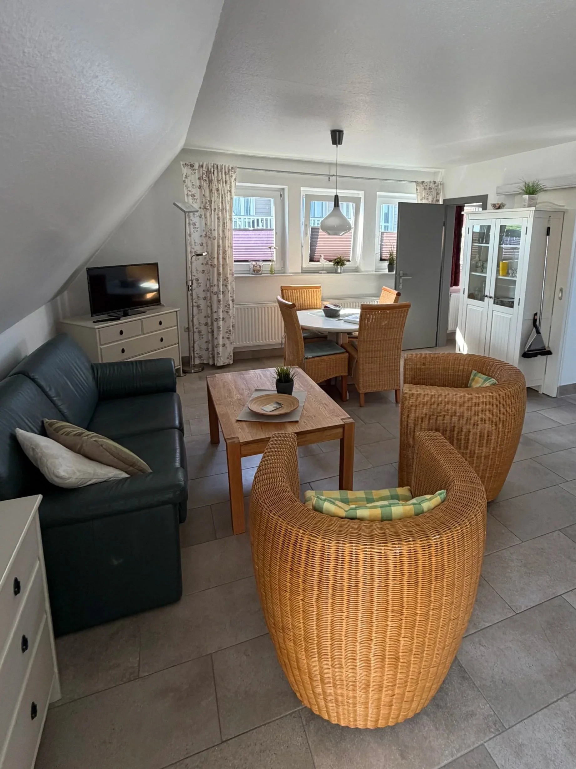Ein Wohnzimmer mit einem Esstisch, sechs Korbsesseln, einem schwarzen Sofa, einem Fernseher, einem weißen Sideboard, Fenstern mit Blumenmuster-Vorhängen und einer vitrine.