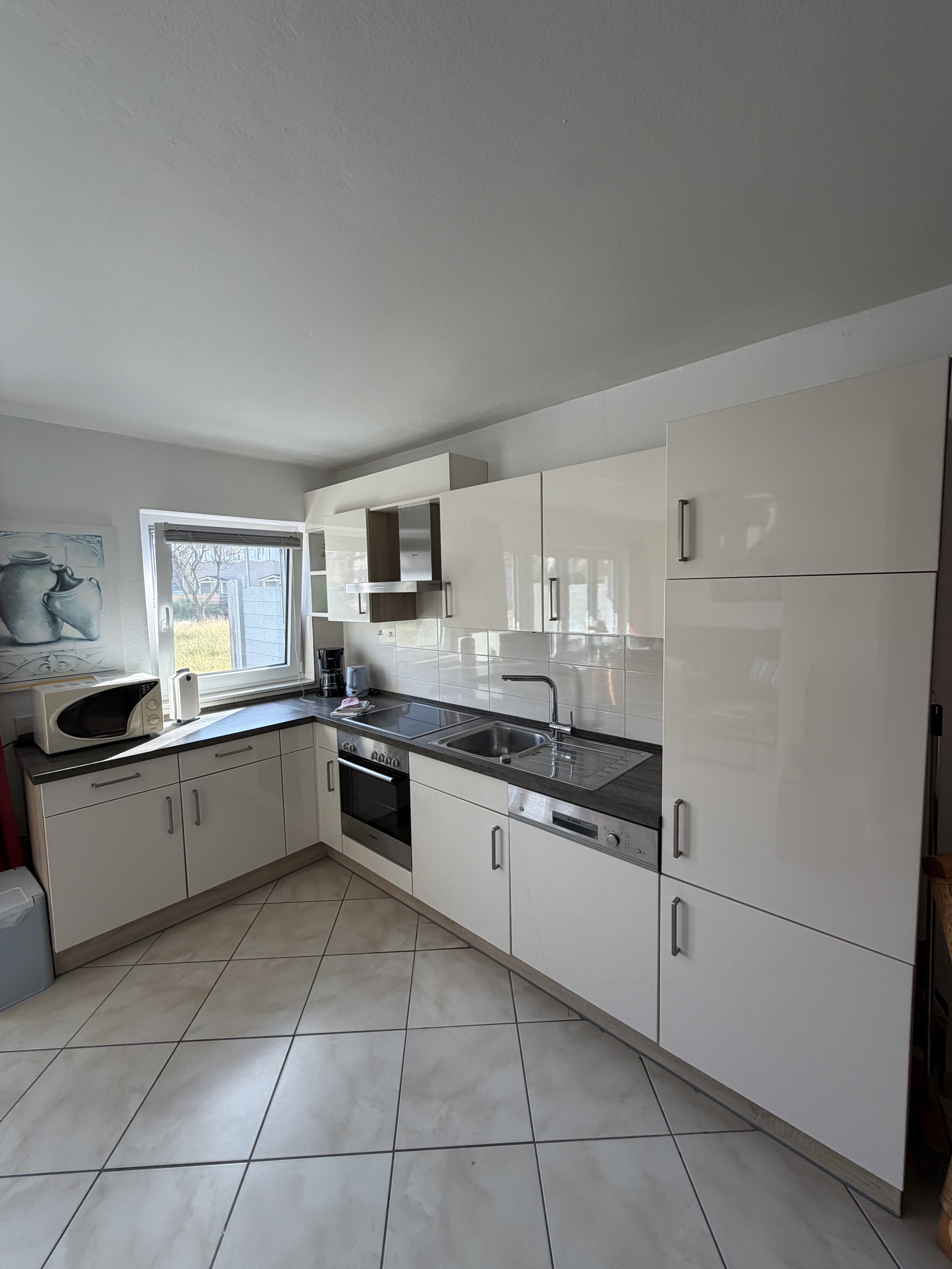 Moderne Küche mit weißen Schränken, Kühlschrank, Herd und Spüle, Sonnenlicht durch Fenster, Kachelboden.