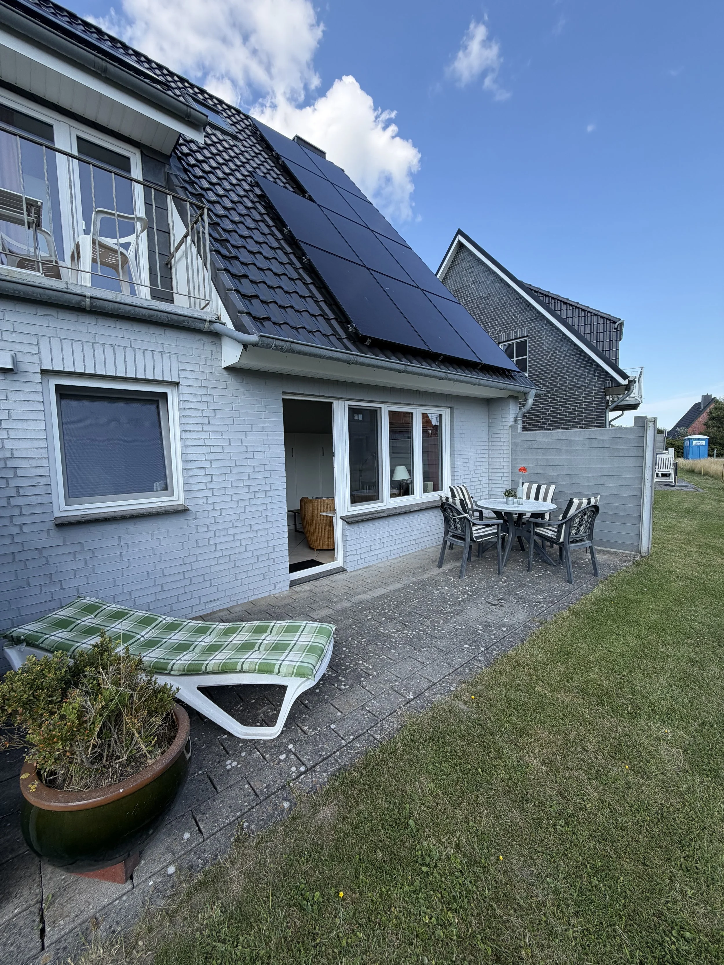 Ein Haus mit Photovoltaik-Anlagen auf dem Dach, ein Balkon mit Stühlen und Tischen, ein Garten mit Rasen und Pflanzen.