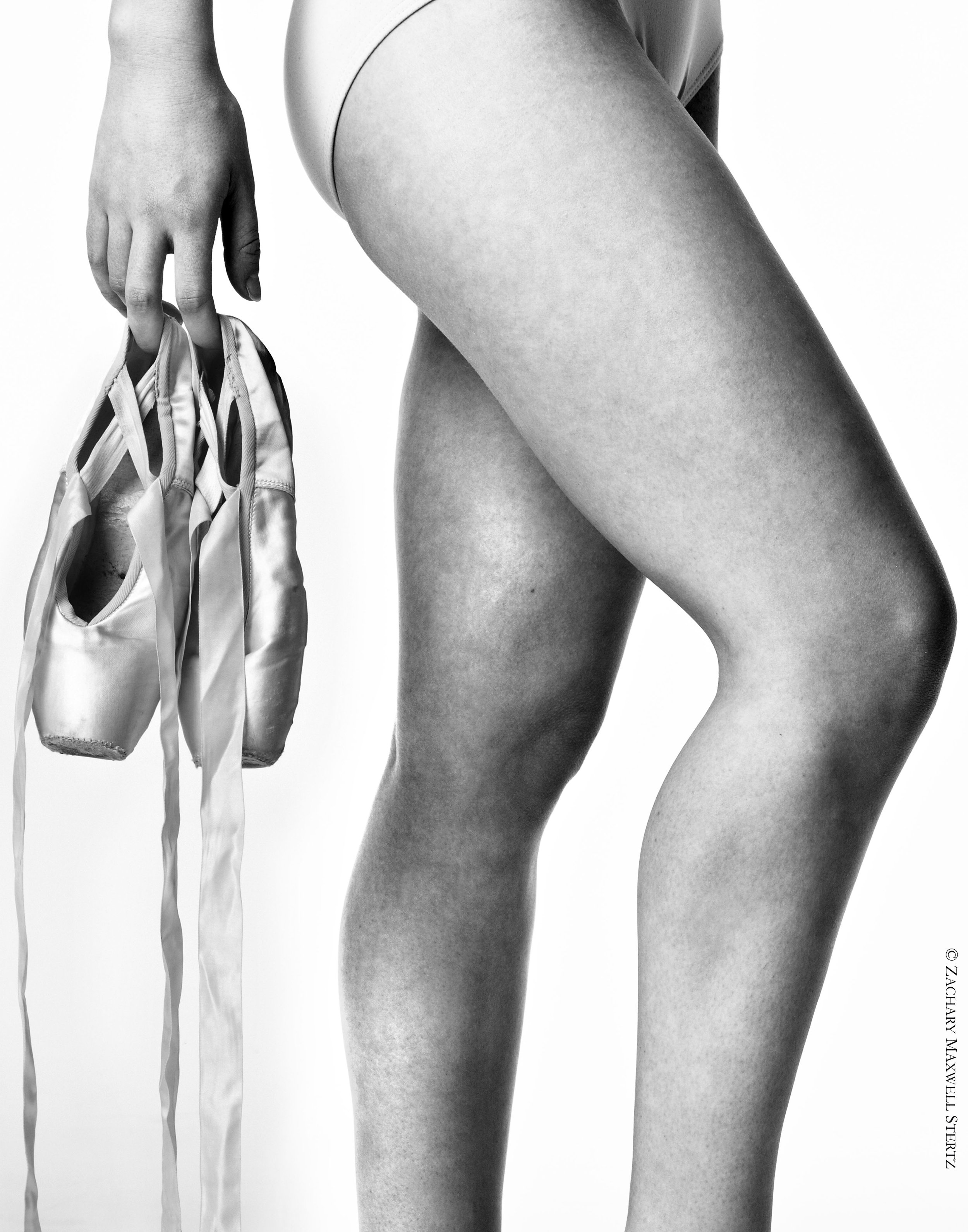Close-up black and white photograph of a dancer's legs, with one hand holding a pair of pointe shoes