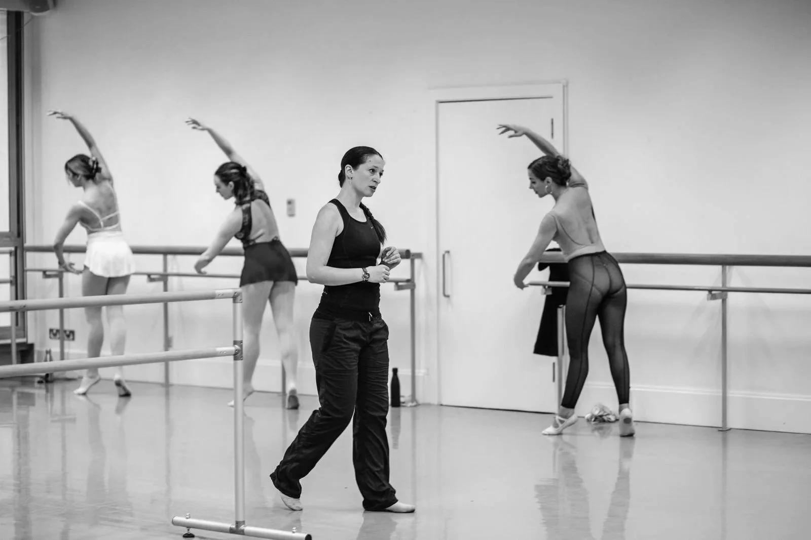 Ballet class in progress with four women, one instructor and three students, in a dance studio.