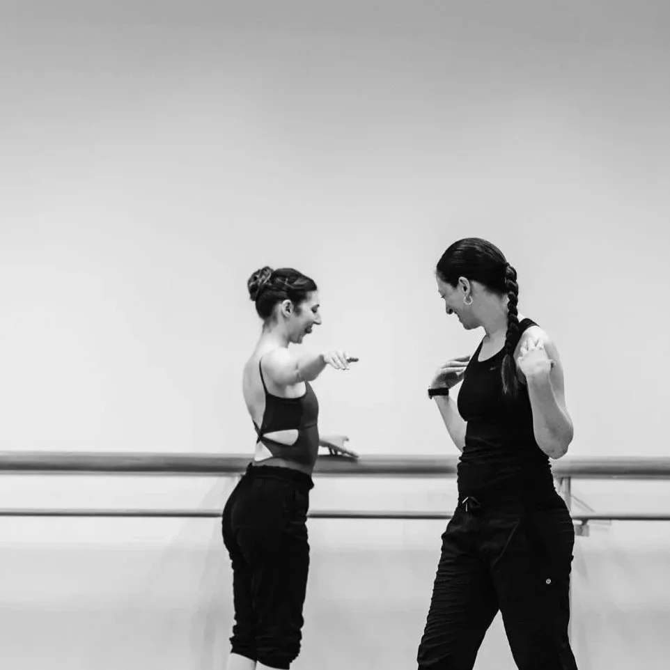 Two women in workout attire laughing and having fun during a dance or fitness class in a studio with a ballet barre.