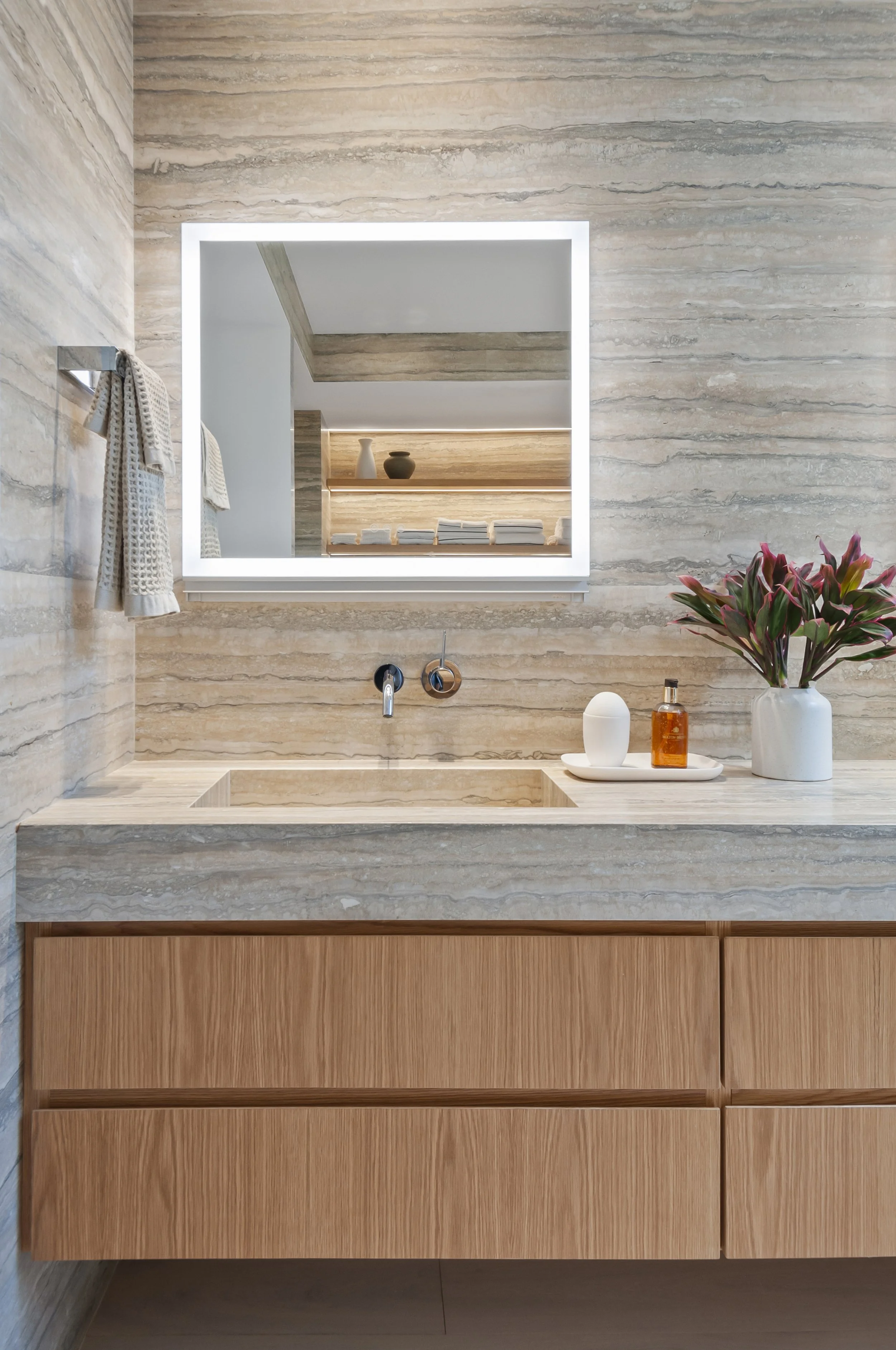 Bathroom vanity with beige marble countertop, a rectangular undermount sink, a large illuminated mirror, a potted plant with pink and green leaves, and various toiletries on the tray.