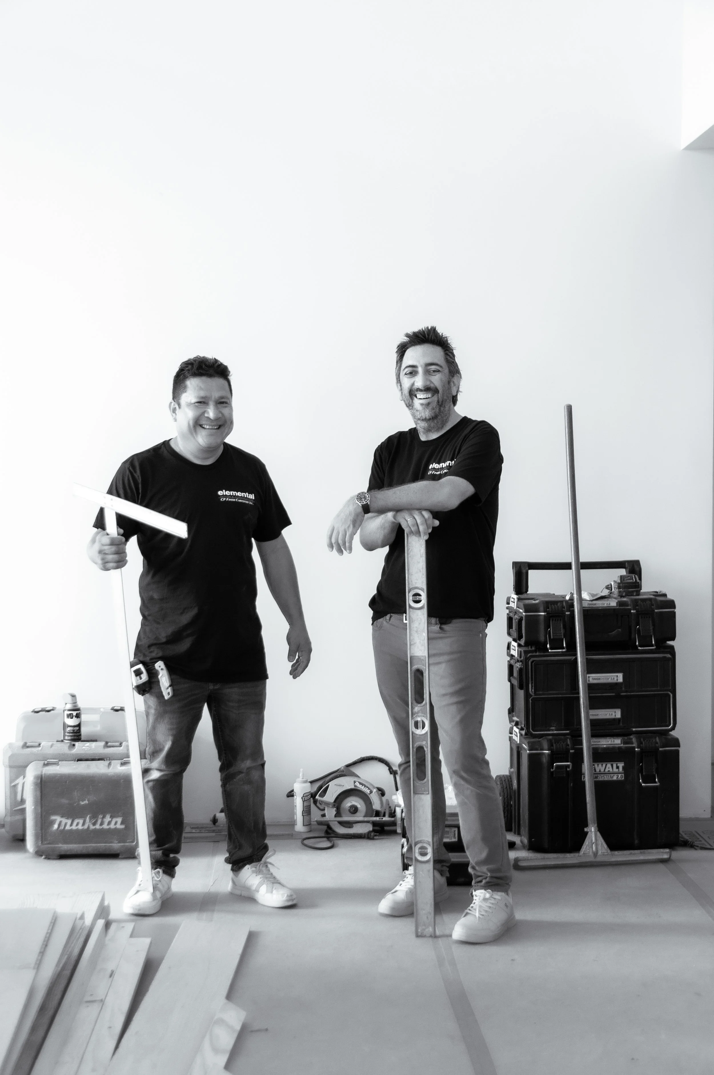 Two men standing in a room with construction tools and equipment. One man is smiling and holding a measuring square, the other has his arms crossed and is leaning on a level. The room has a plain white wall and various power tools and boxes are visible around them.